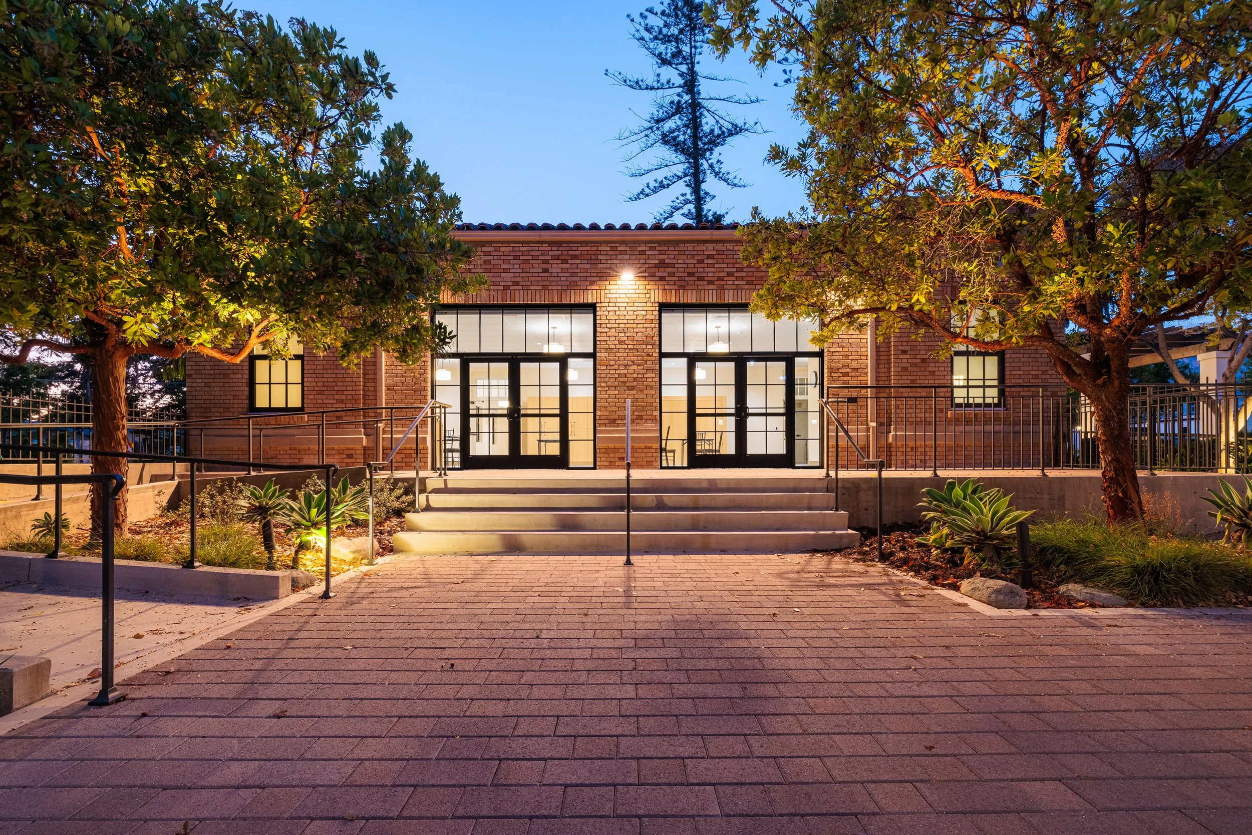 A brick building with two large glass doors and windows, surrounded by trees and landscaping, illuminated at dusk.