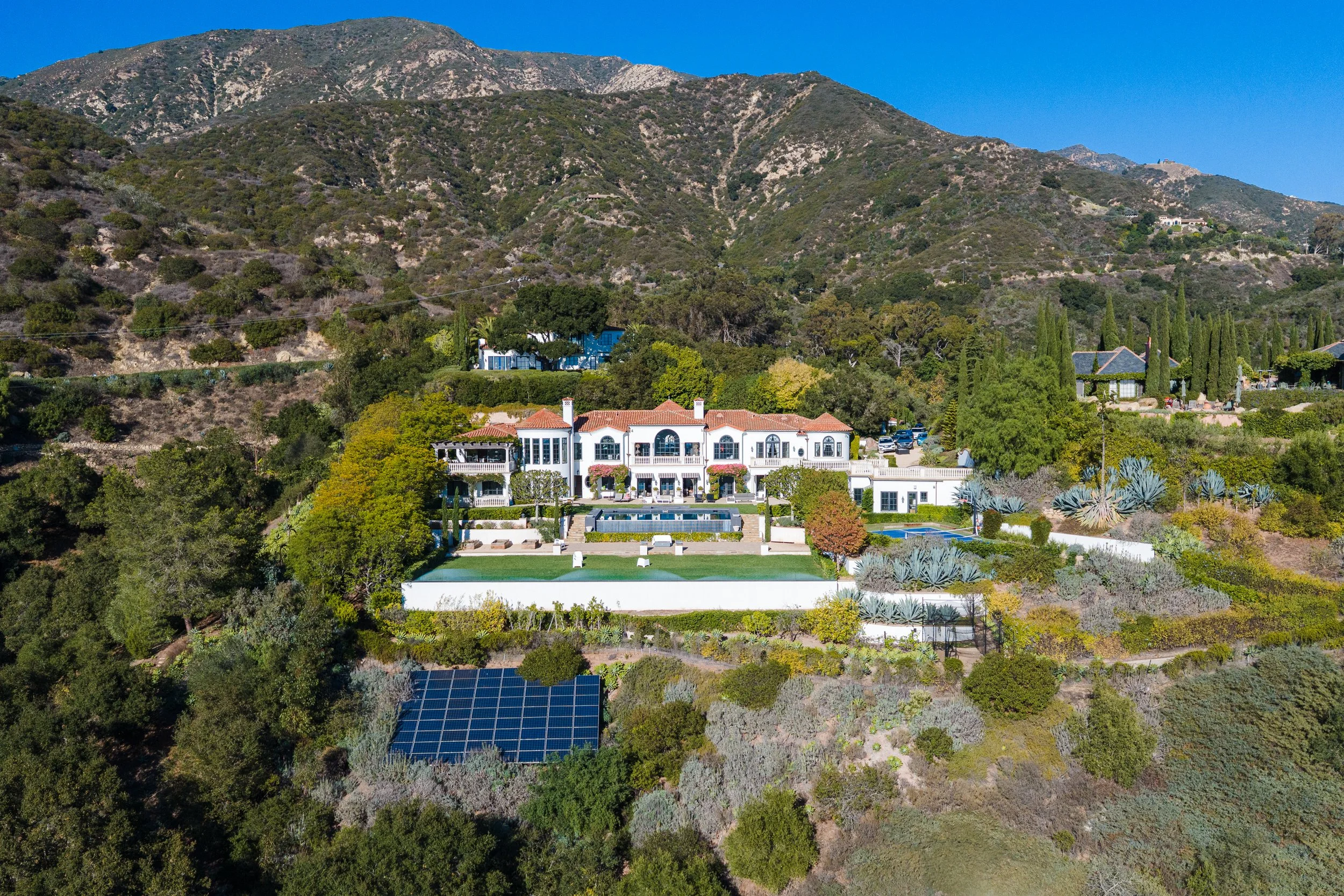 A large, luxurious white mansion with red-tiled roof, multiple terraces, and a swimming pool, located on a hillside surrounded by trees, agave plants, and mountain scenery under a clear blue sky.
