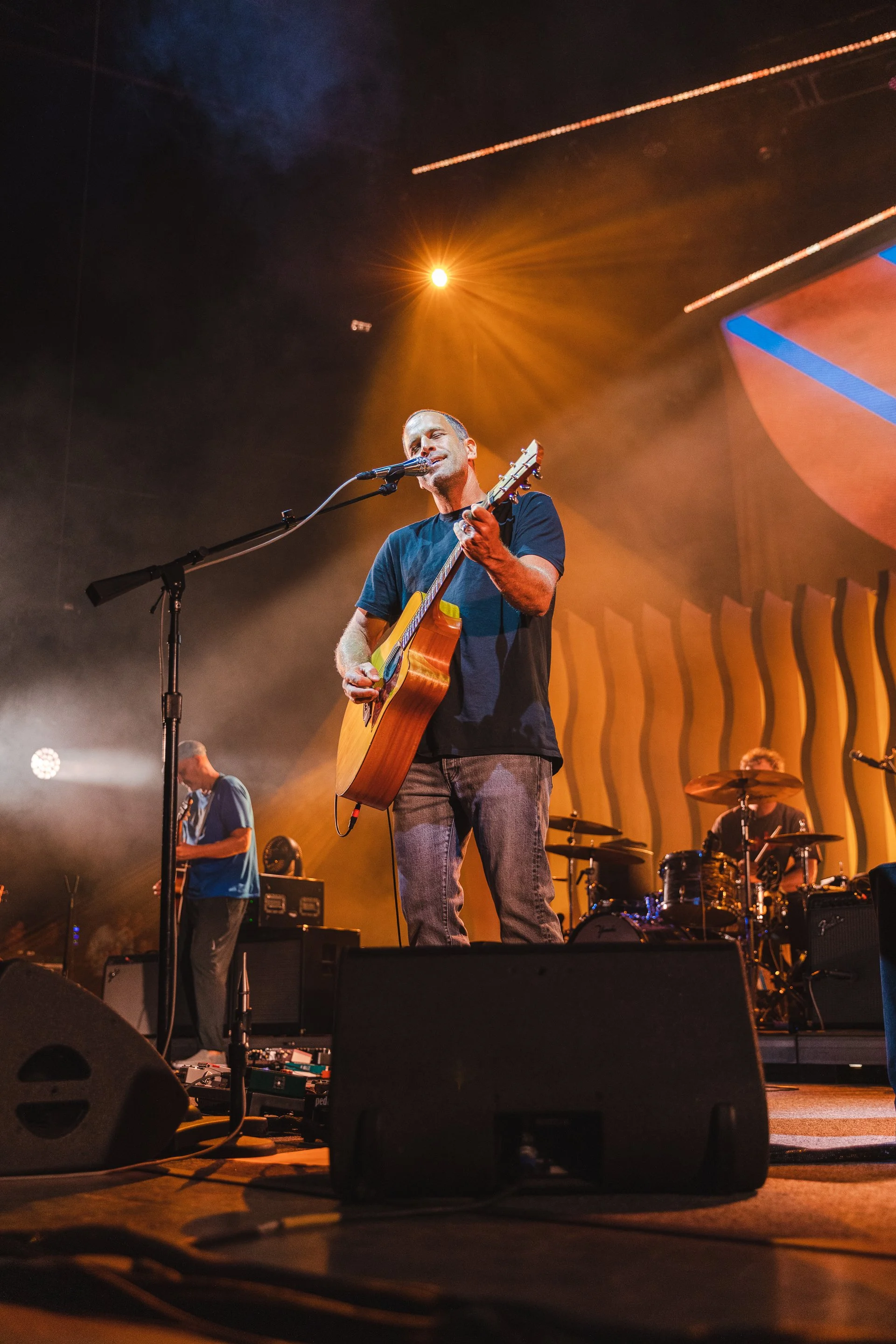 Jack Johnson playing an acoustic guitar and singing into a microphone on stage with band members and vibrant lighting.