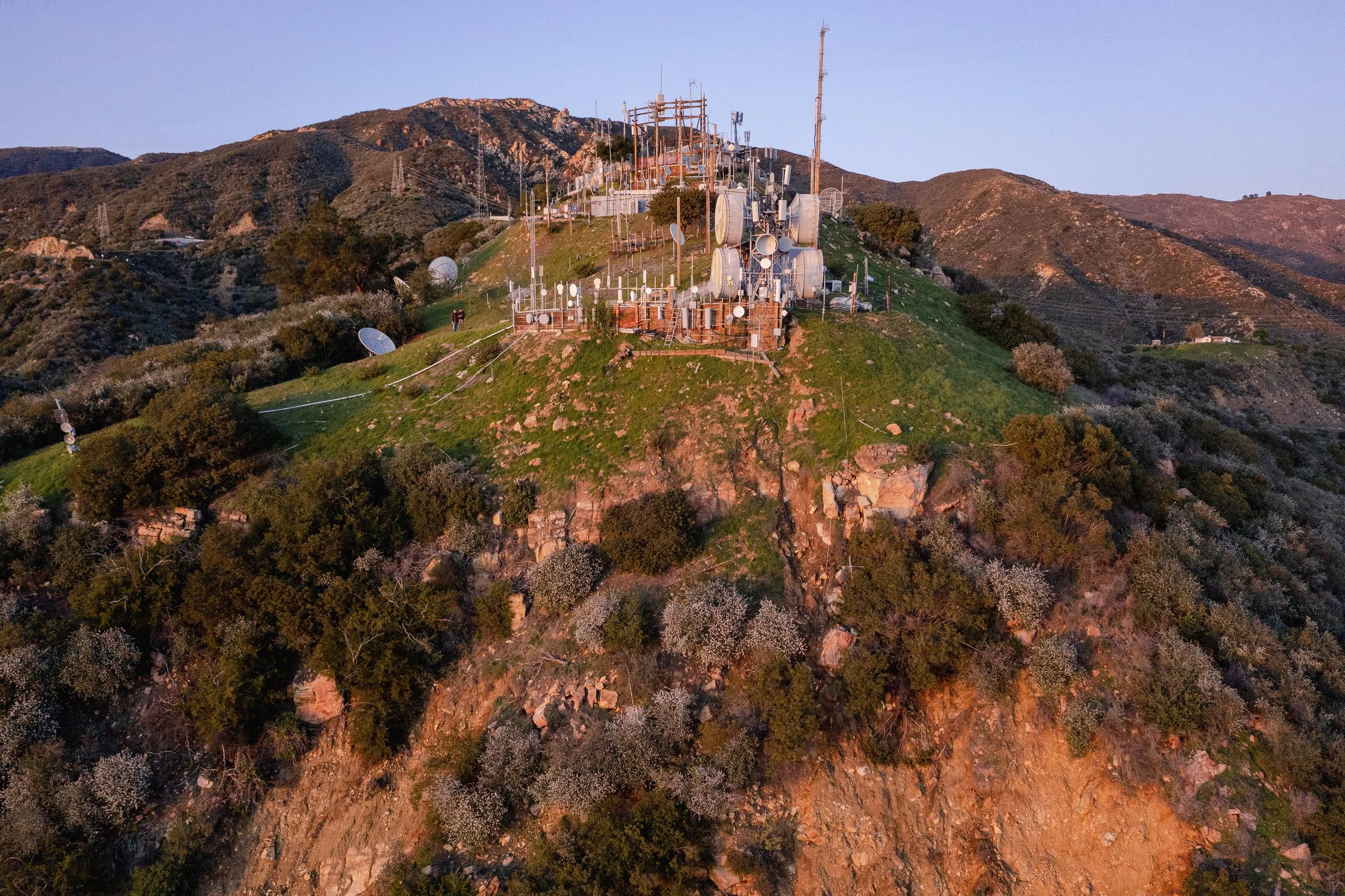 A hilltop area with numerous communication towers and antennas, surrounded by trees and shrubs, with mountains in the background.