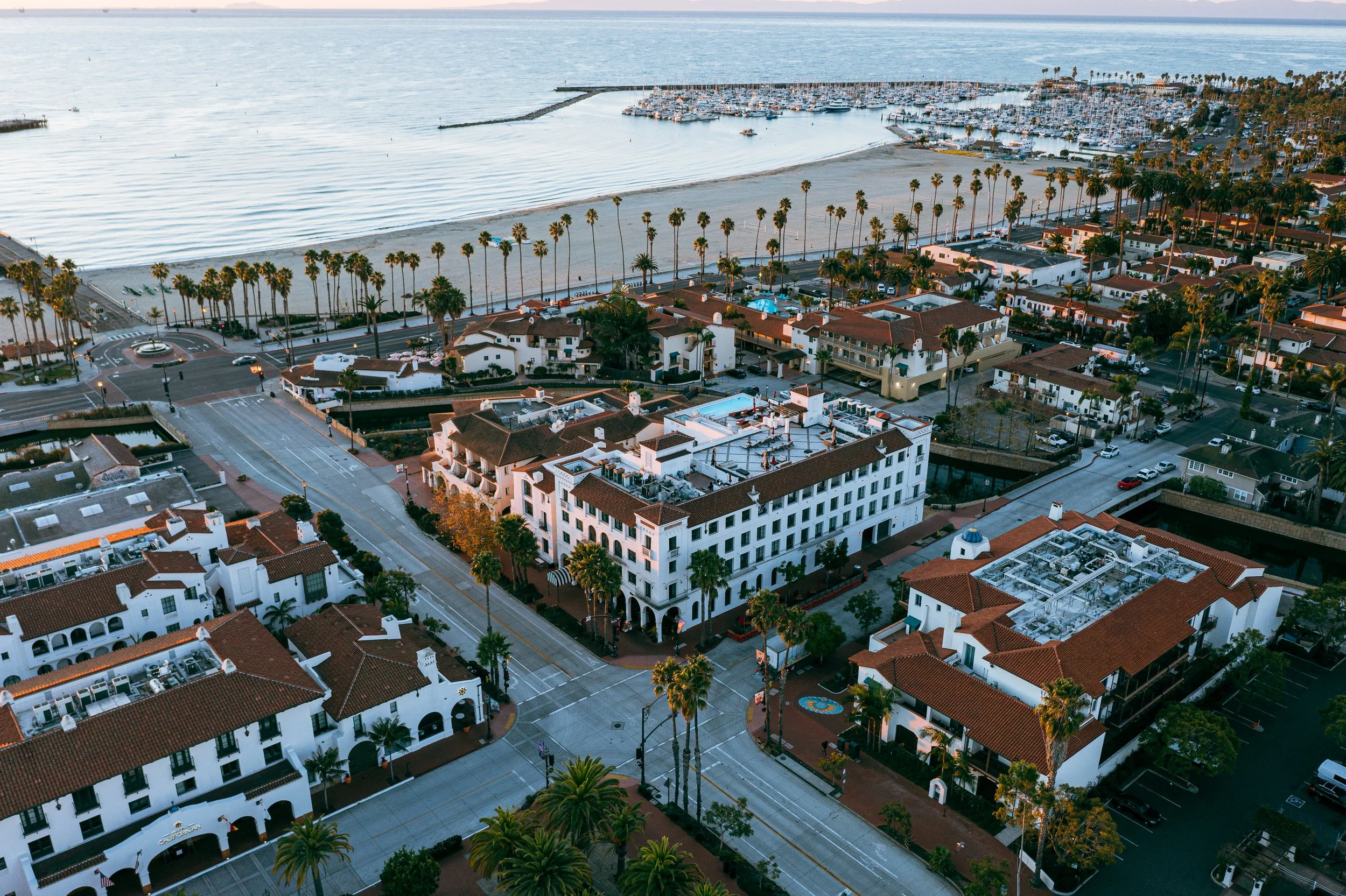 Aerial view of a coastal town showing white buildings with red-tile roofs, palm trees lining the streets, and a beach with a marina filled with boats in the background.
