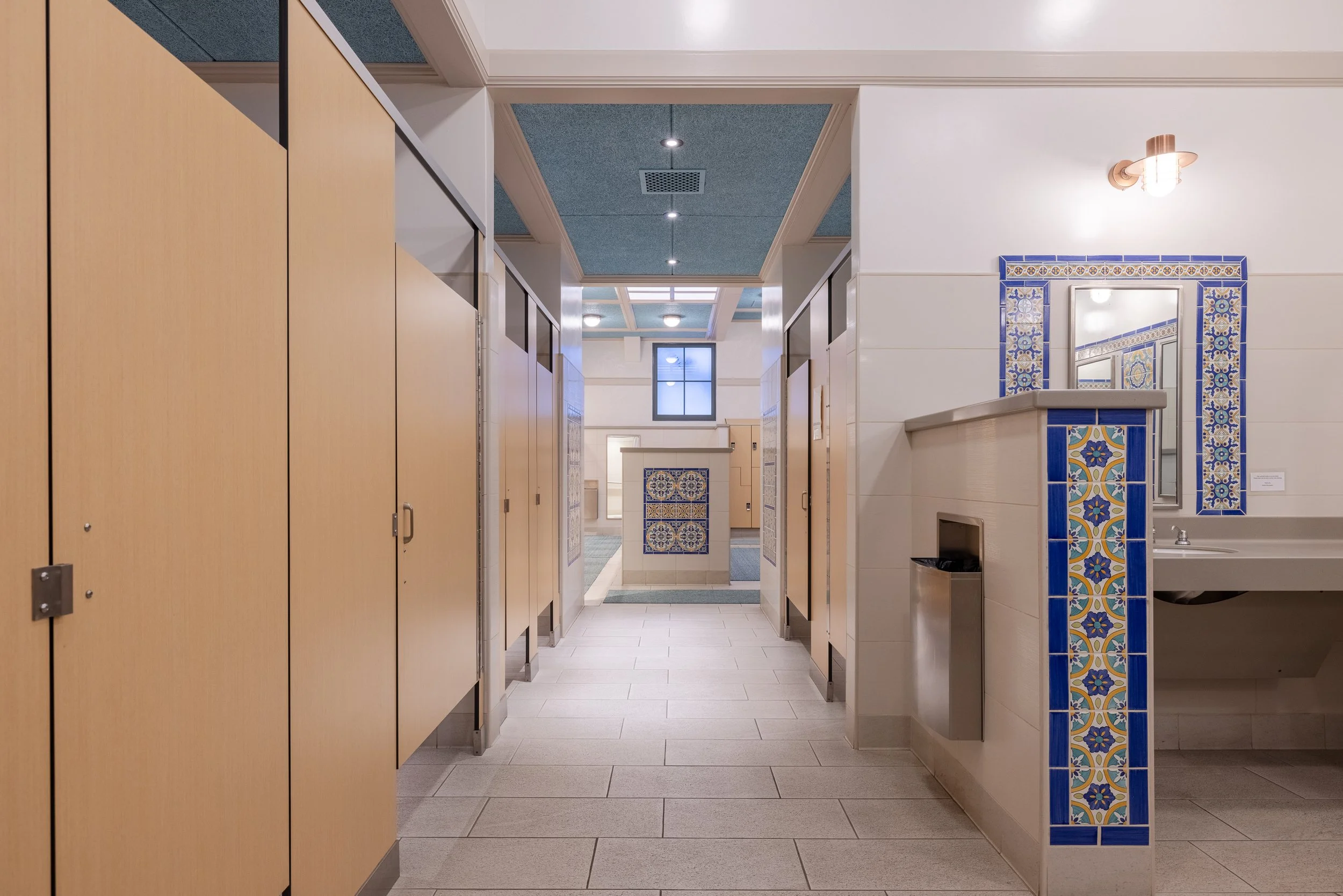 Public restroom with beige stalls, tiled walls with blue and yellow decorative patterns, a mirror above the sink, and a trash bin on the floor.