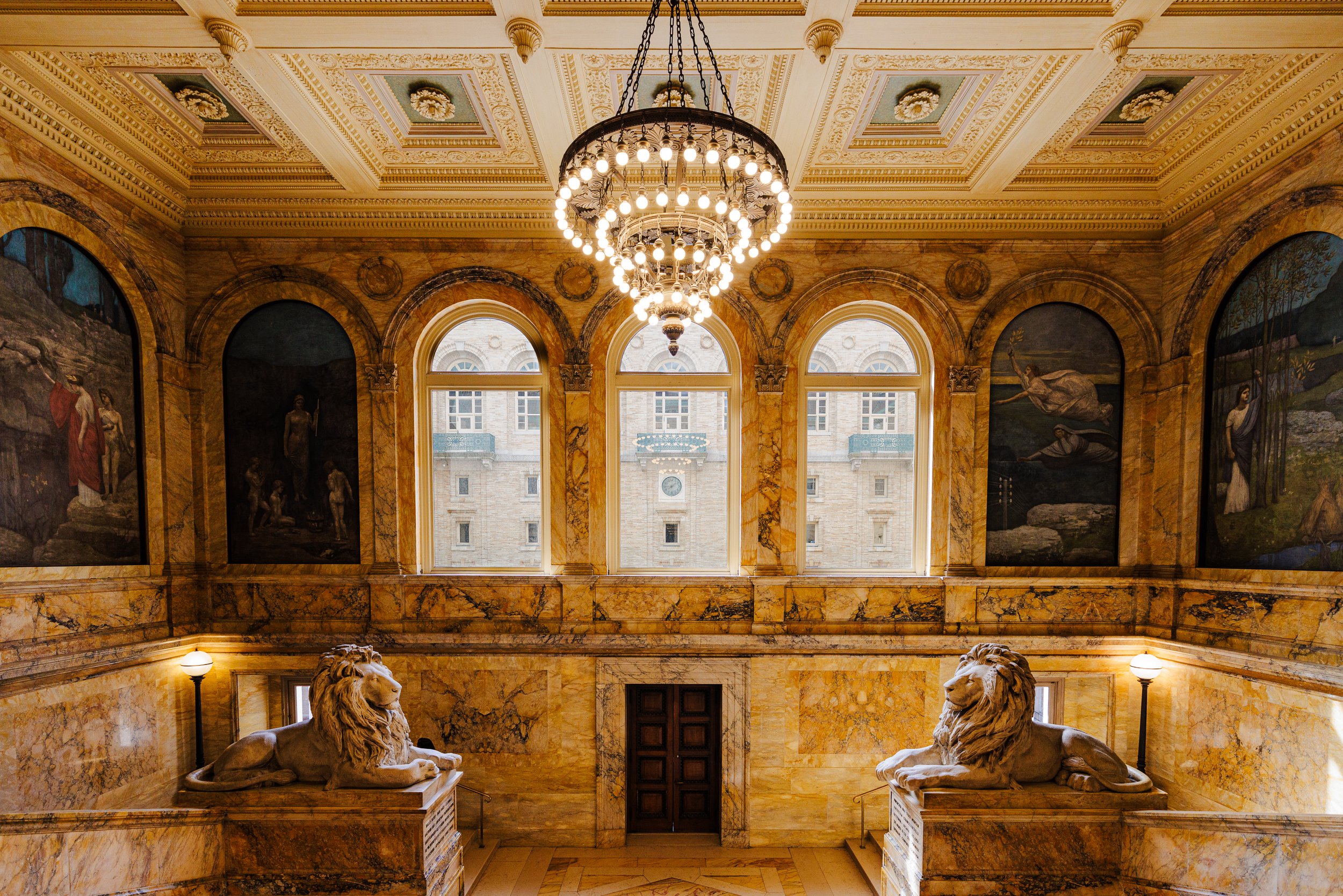 Interior of a grand building with ornate marble walls, a chandelier hanging from a decorated ceiling, and two large stone lion statues at the staircase landing. There are three large arched windows letting in natural light and paintings on the walls.