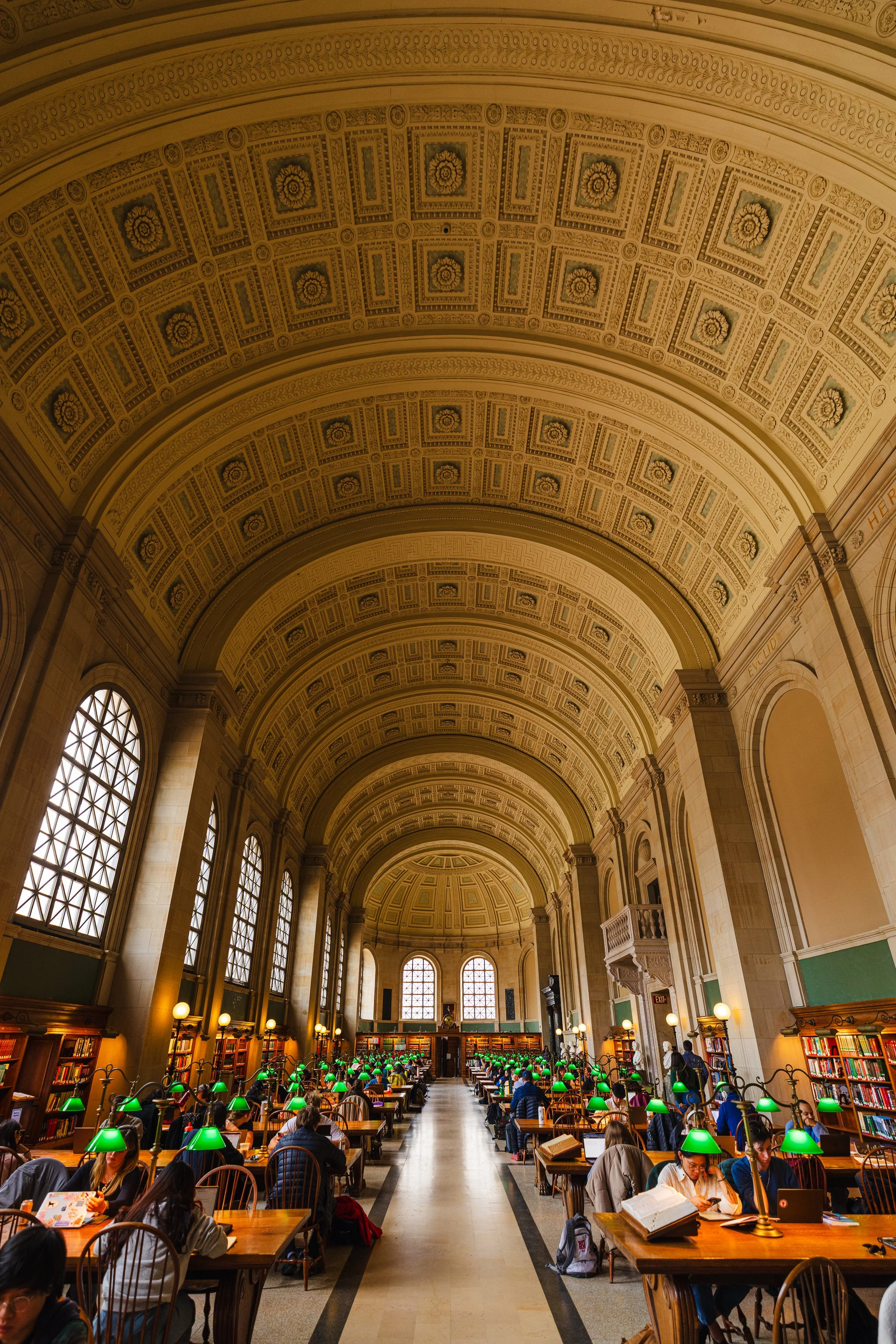 A grand, historic library with high vaulted, decorated ceilings and large arched windows. Numerous people seated at wooden desks under green reading lamps, studying and reading.