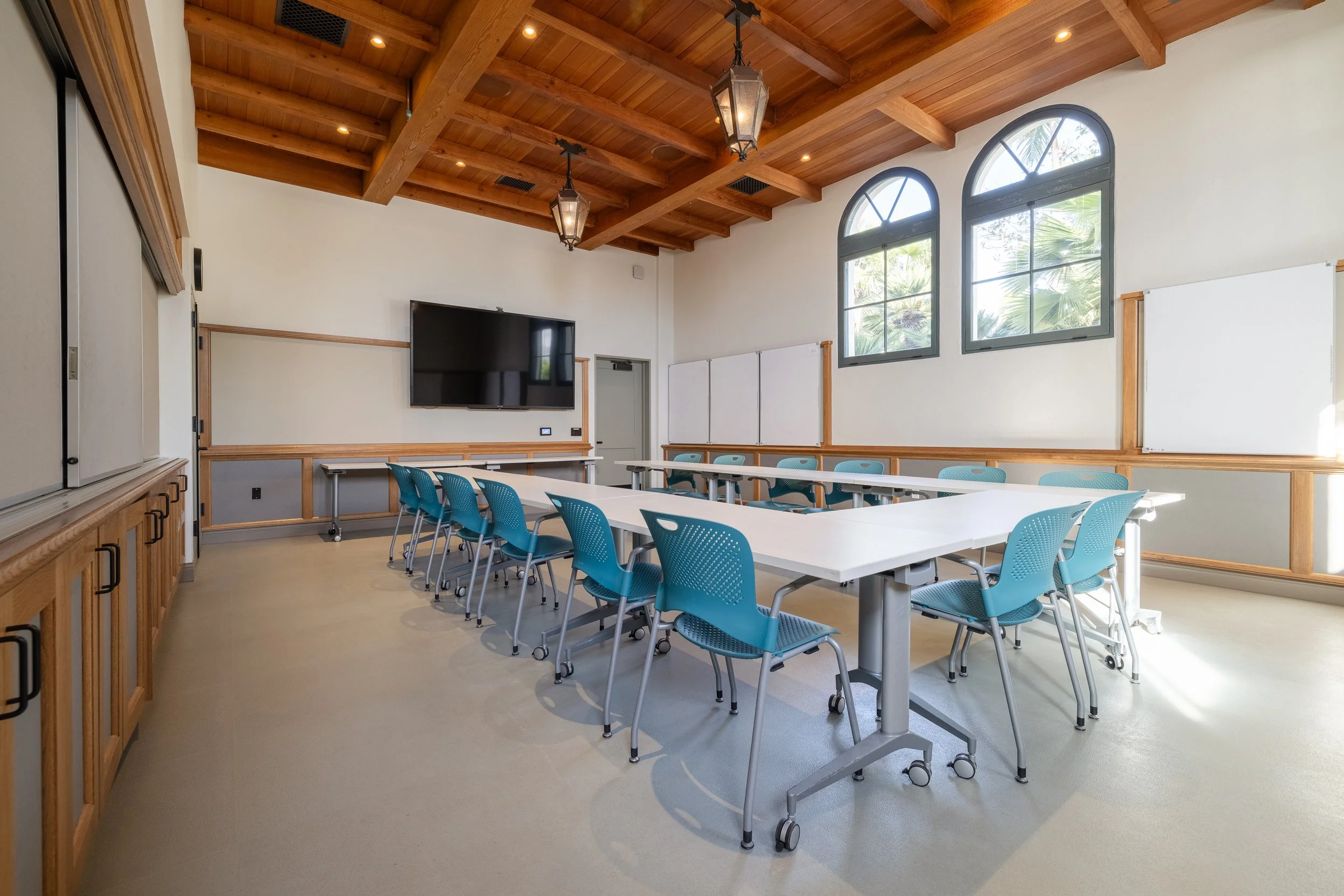 A modern conference room with a wooden ceiling, blue chairs around white tables, large arched windows, and a flat-screen TV on the wall.
