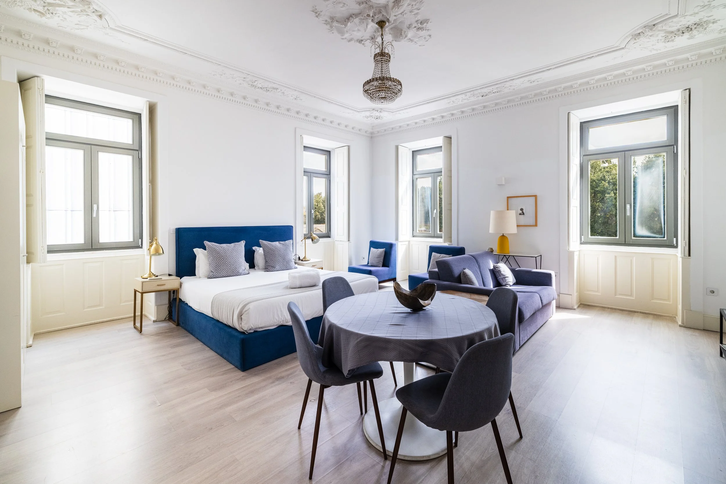 Bright, spacious living room with white walls, large windows, and modern furniture including a blue bed, blue chairs, a purple sofa, and a dining table with six chairs. There are gold and yellow accents, a chandelier, and decorative pillows.