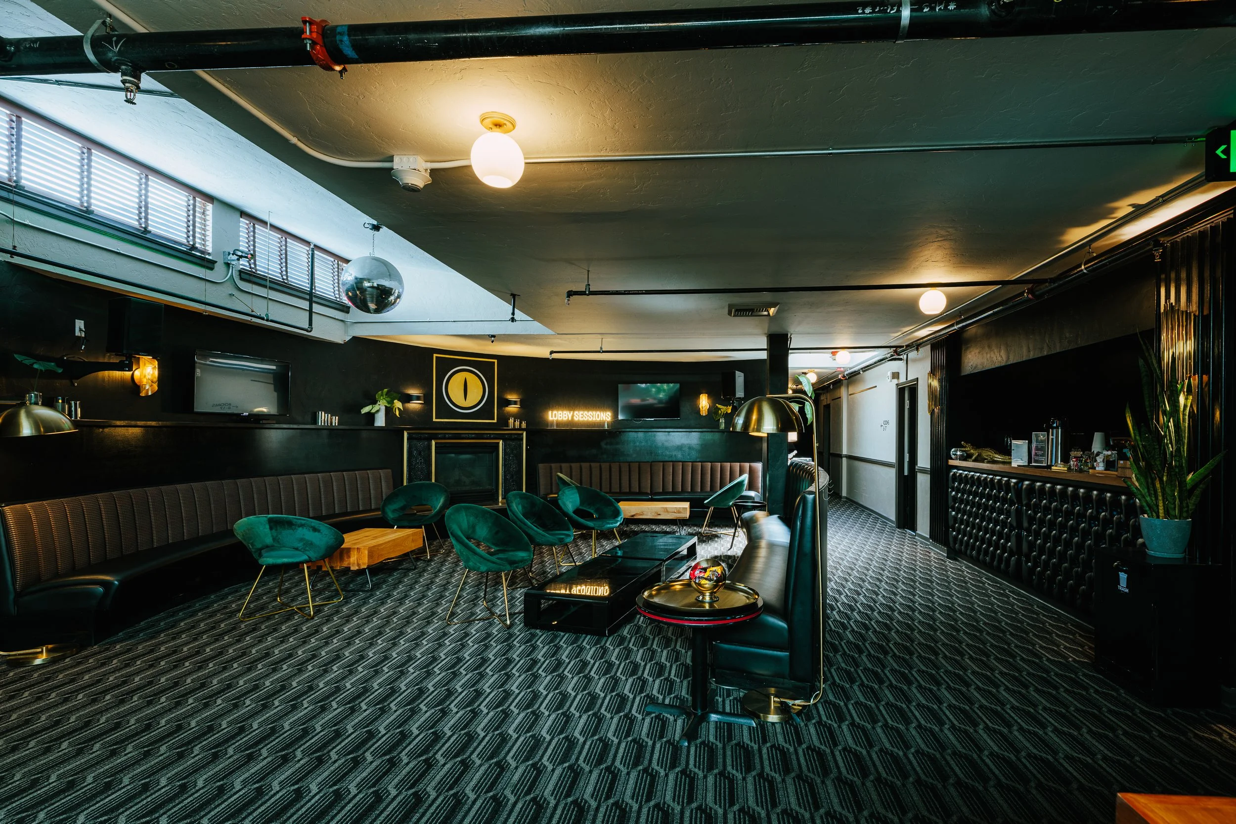 Interior of a lounge with dark walls, modern furniture including green chairs, a black leather couch, and a curved booth. There is a television, a sign that reads 'Lobby Sessions,' a fireplace, and decorative plants. The ceiling has lights, a disco b