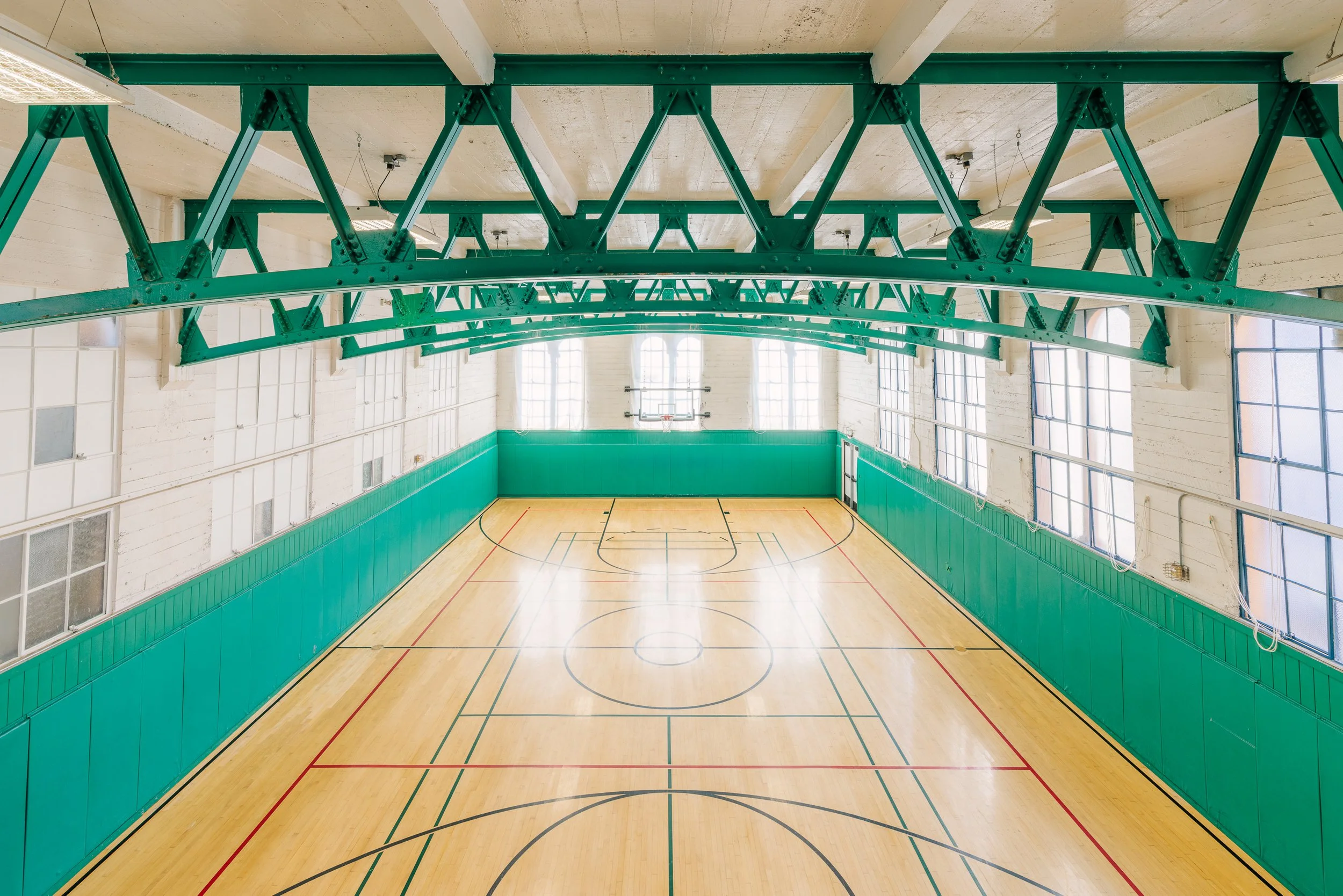 Empty indoor basketball court with green and white walls, wooden floor, and large windows, viewed from above.