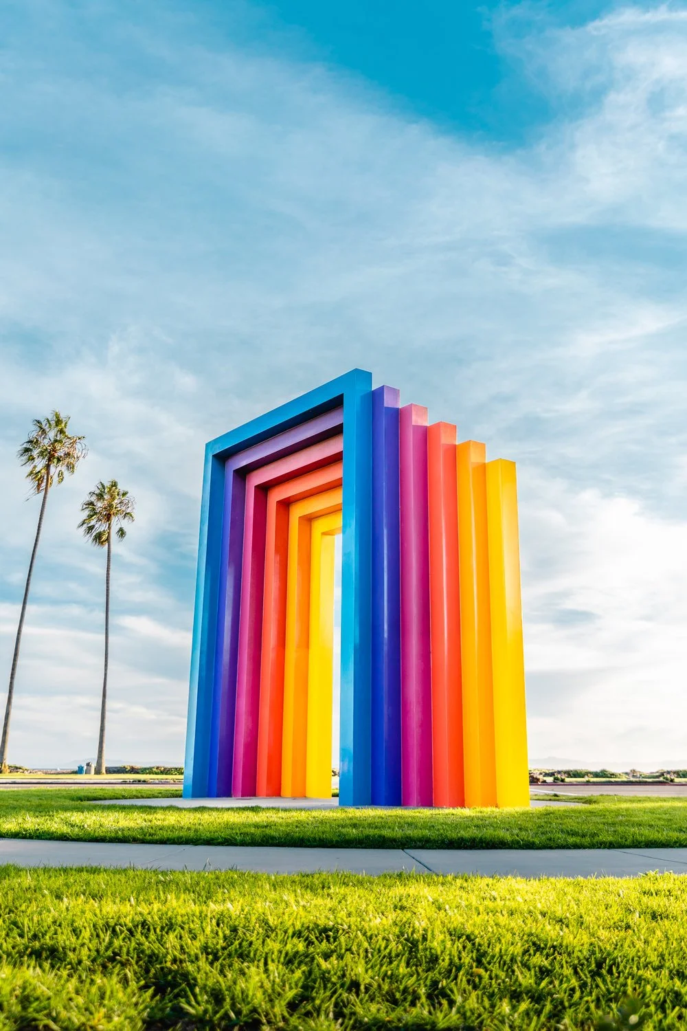 Colorful rainbow-colored art installation with vertical rectangular frames in a park with green grass, palm trees, and cloudy sky.
