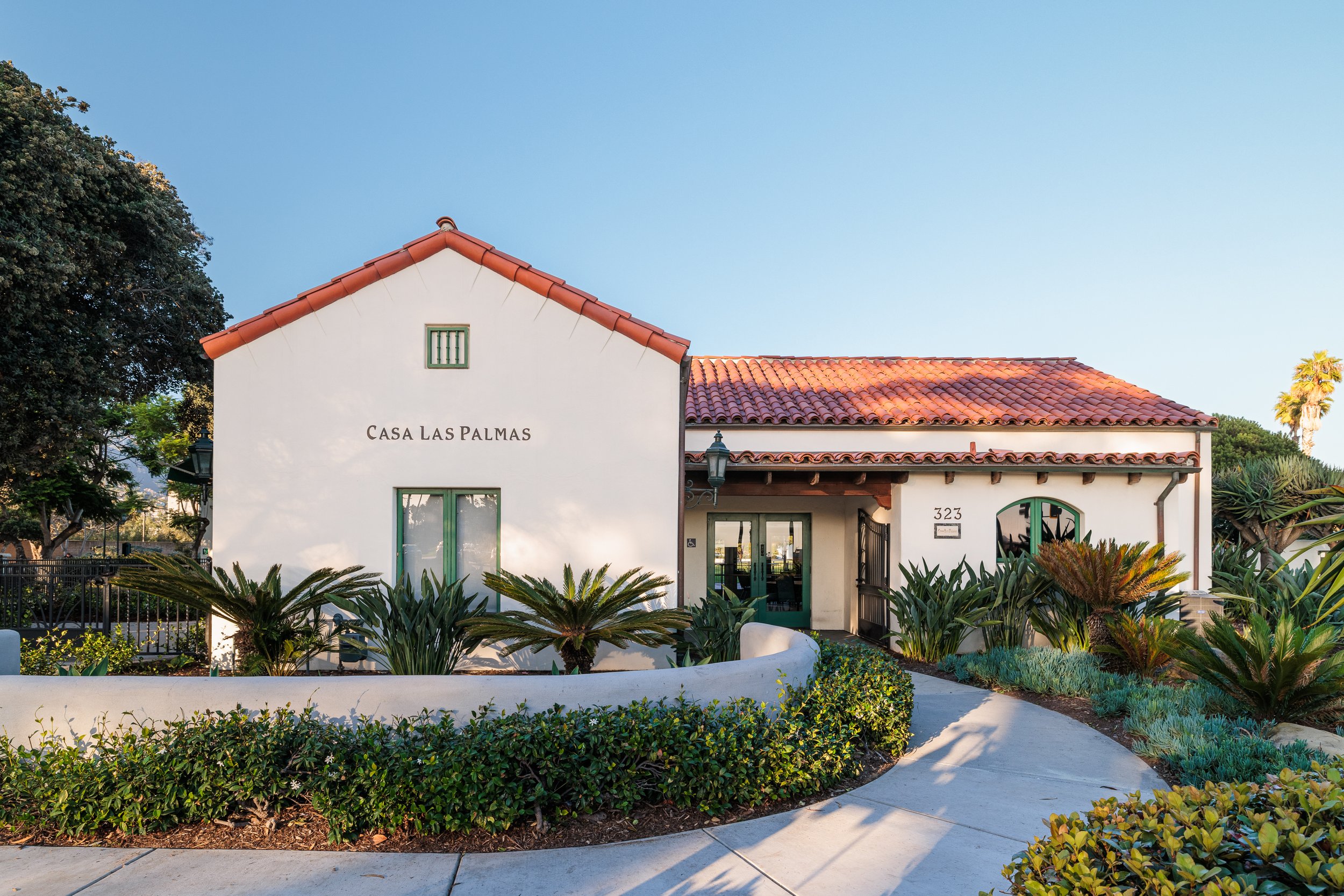 A white building with a red tile roof and green accents, labeled 'Casa Las Palmas', surrounded by lush greenery and a curved sidewalk leading to the entrance, under a clear blue sky.