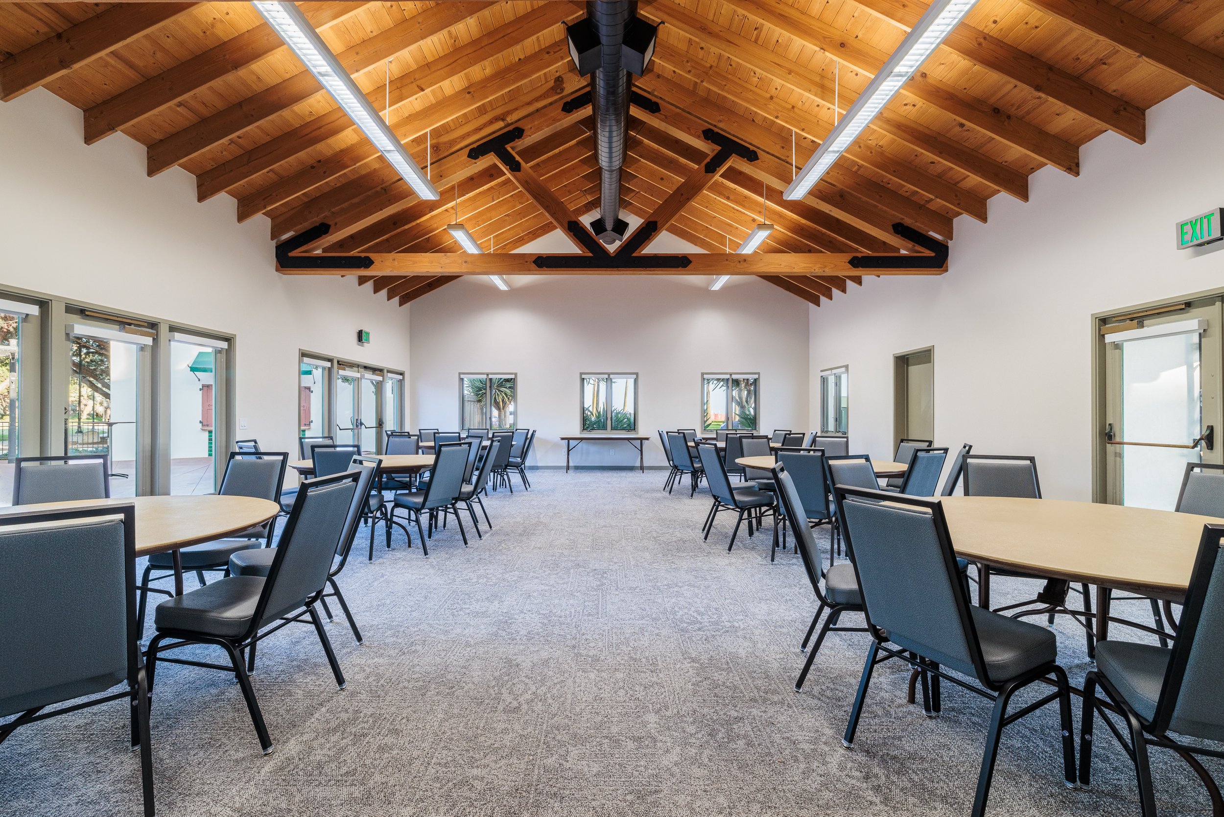 Empty banquet or conference hall with wooden ceiling, round tables with chairs, windows and glass doors along the sides, and a table at the front.