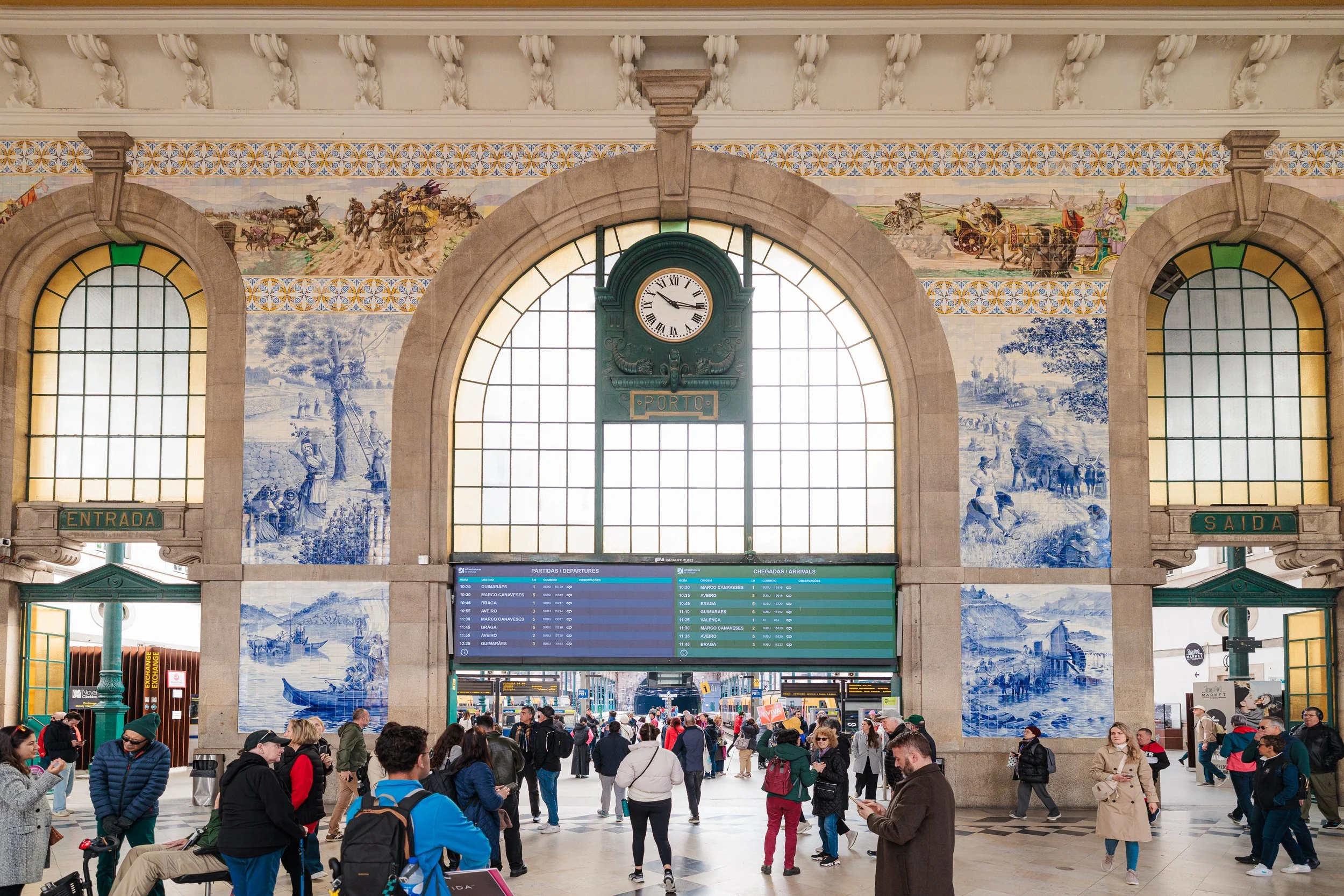 Interior of a busy train station with a large arched window, antique clock, and blue and white tile murals depicting historical scenes. People are walking and waiting, with some looking at departure screens.