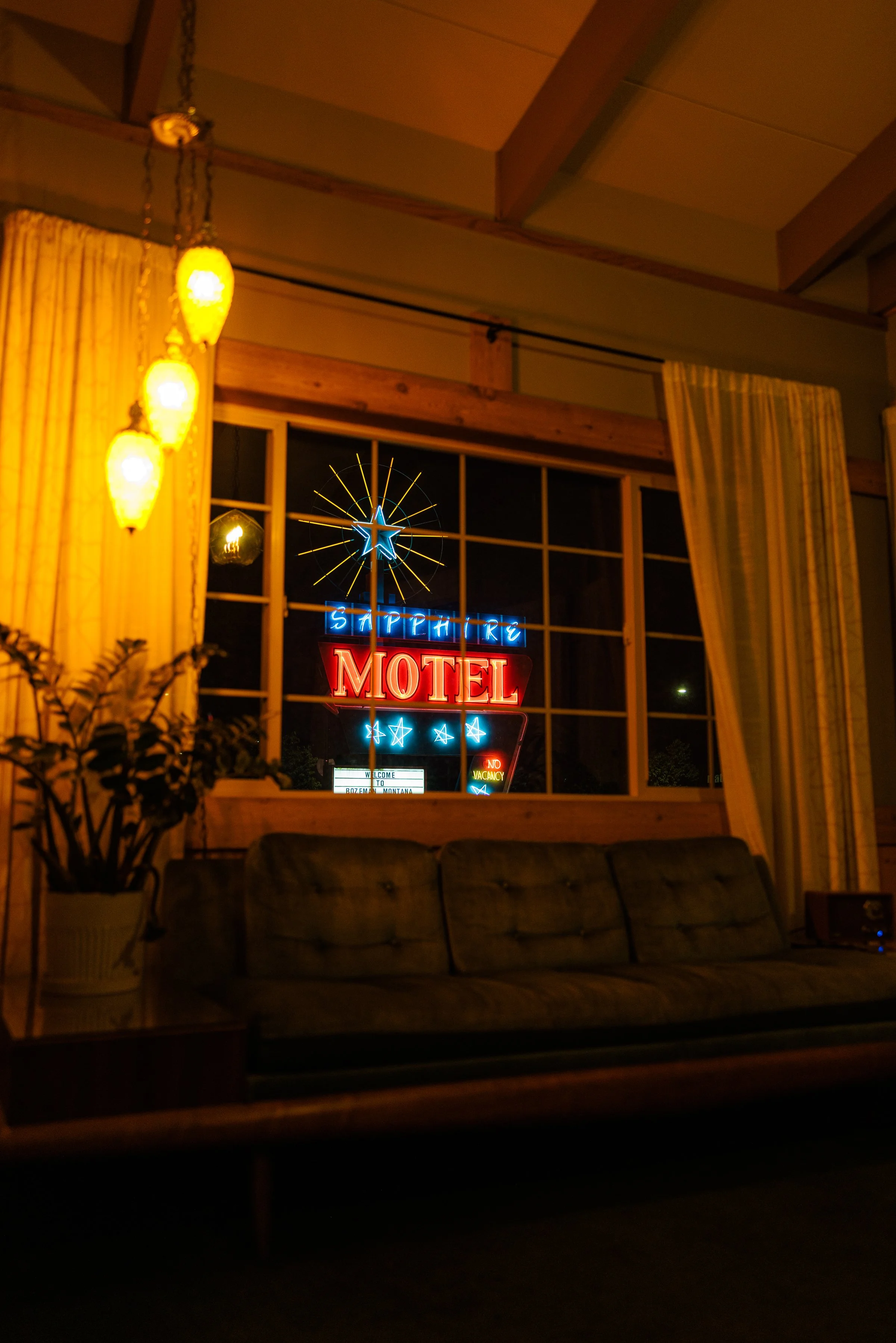 Neon sign outside a motel seen through a window from inside a room with orange curtains, a vintage sofa, and a potted plant.