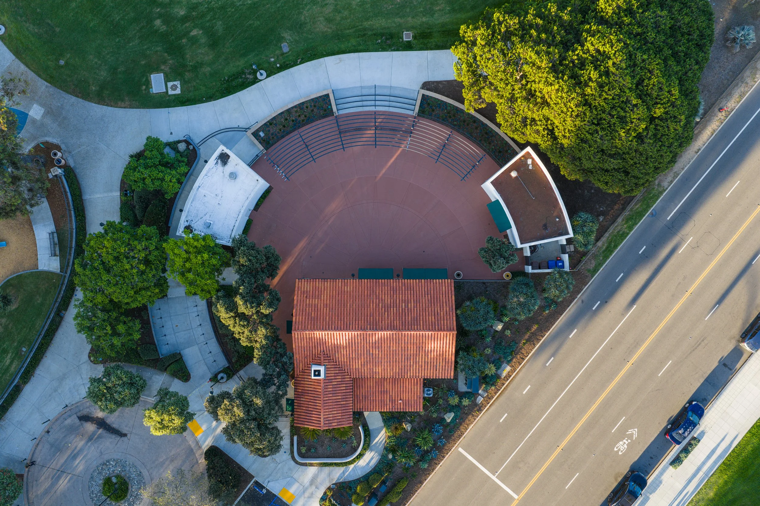 An aerial view of a park area with a large circular stage or amphitheater, surrounded by trees, walkways, and a building with a red tile roof adjacent to the stage. There is a roadway with cars running parallel to the right side of the image.