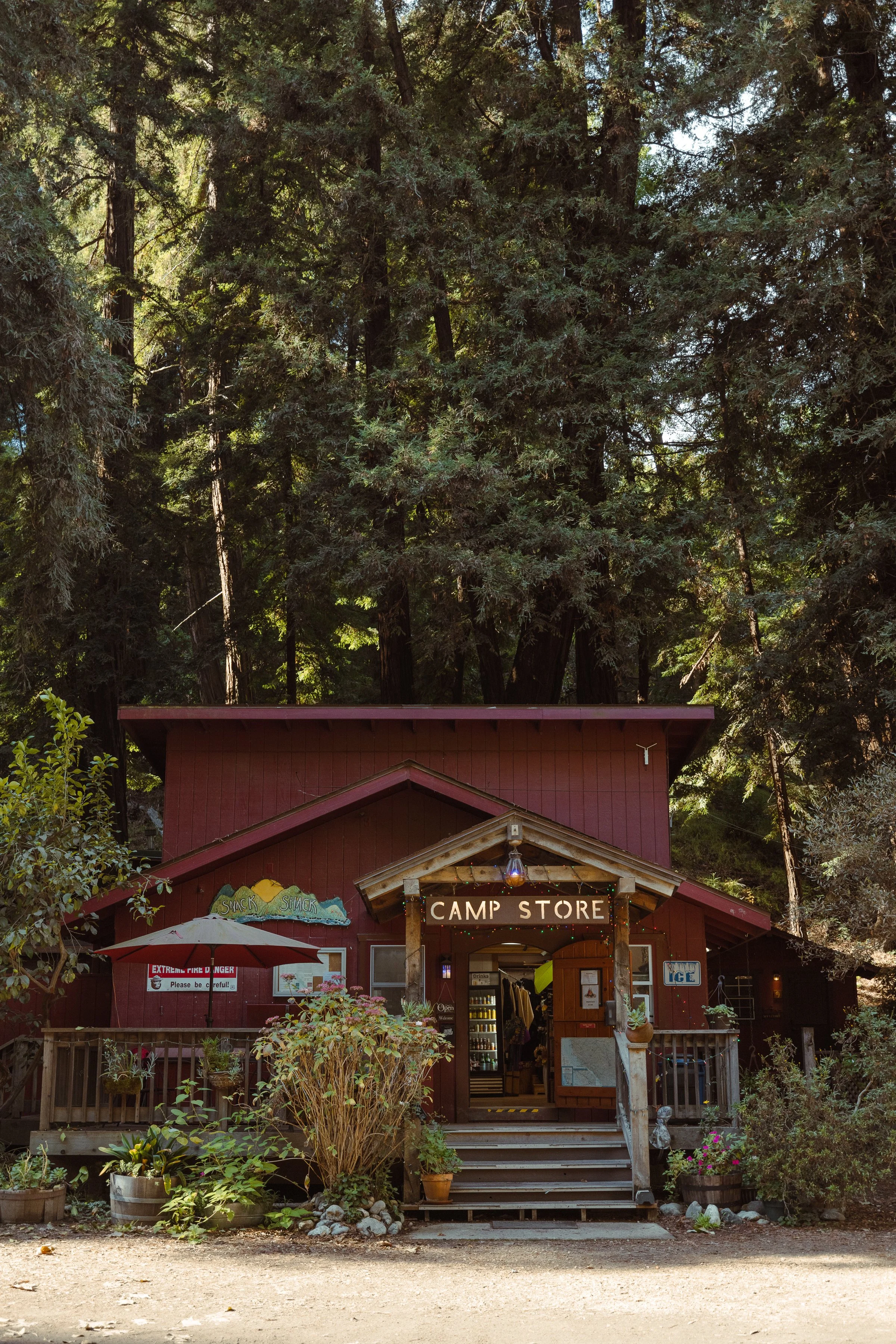 A rustic camp store building with a sign above the entrance, surrounded by trees and plants, with outdoor decor and an umbrella on the left side.