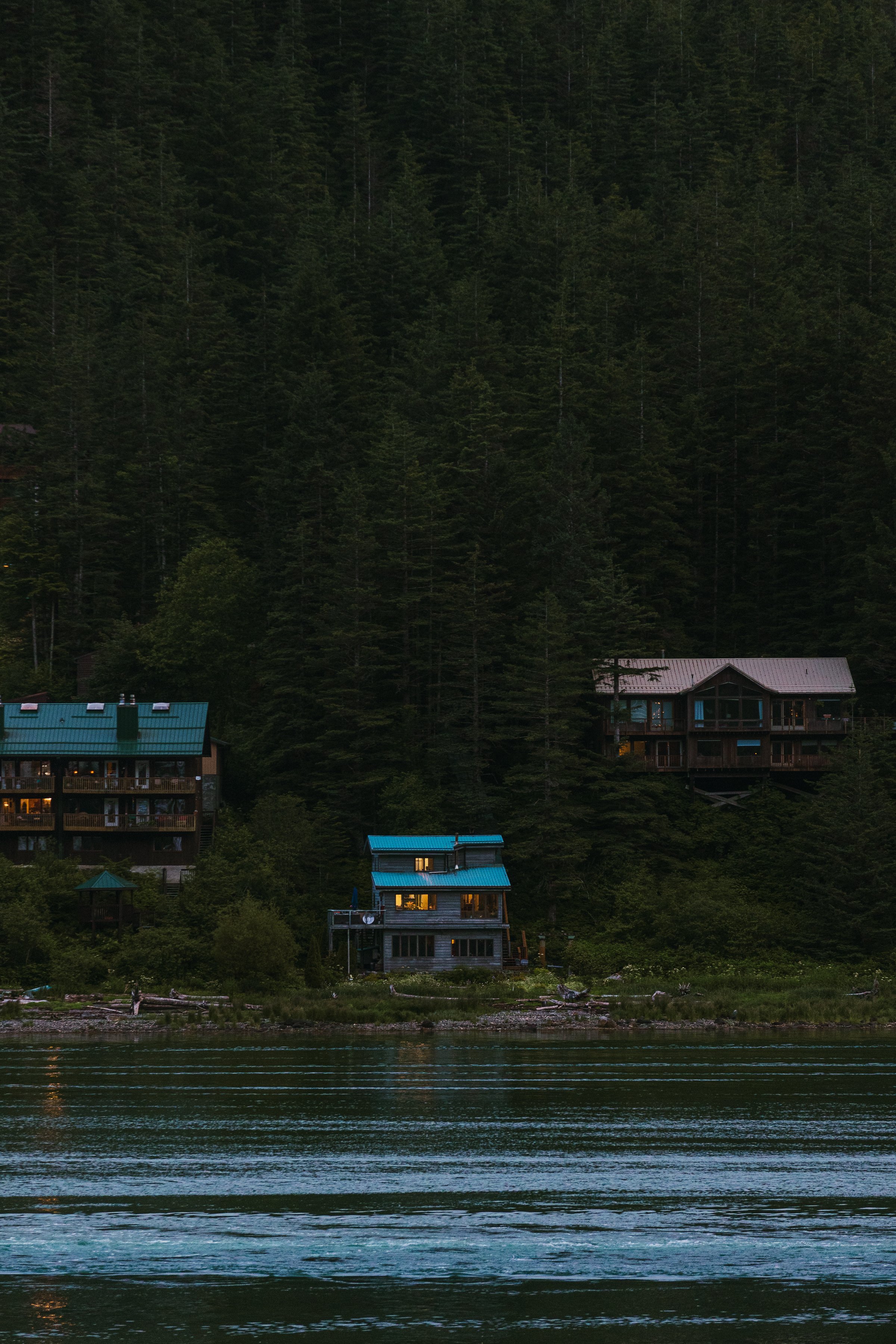 Three houses with blue rooftops situated at the edge of a body of water, surrounded by dense green forest on a hillside at dusk.