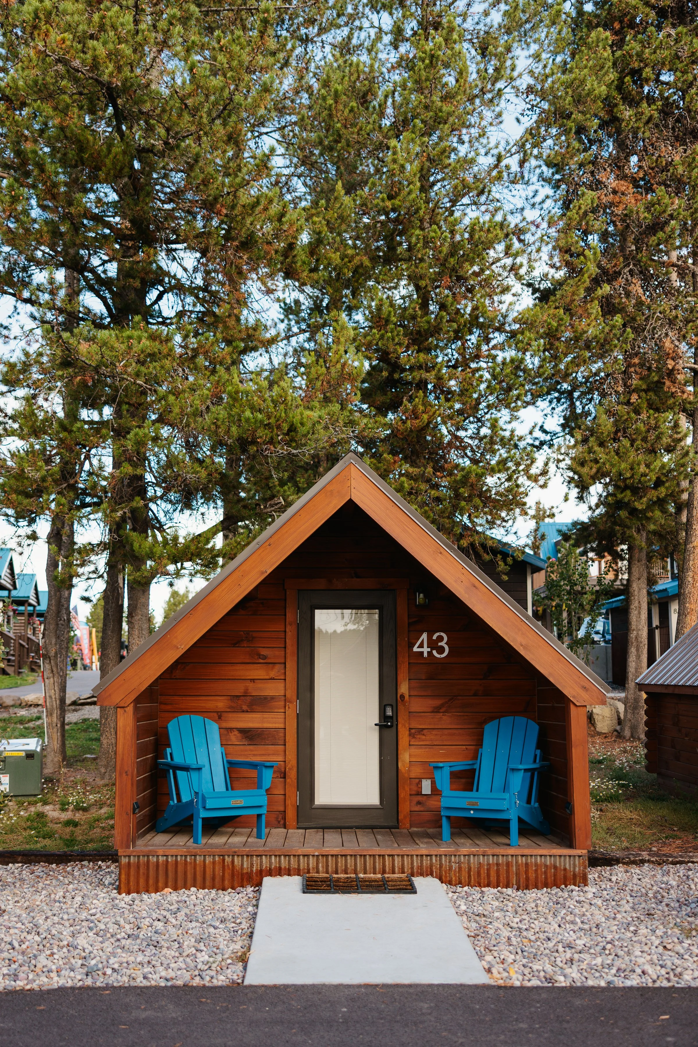 A small wooden cabin with a steep gabled roof, painted in a natural wood tone, with two blue Adirondack chairs on its front porch, and the number 43 displayed next to the door. Tall pine trees surround the cabin in the background.