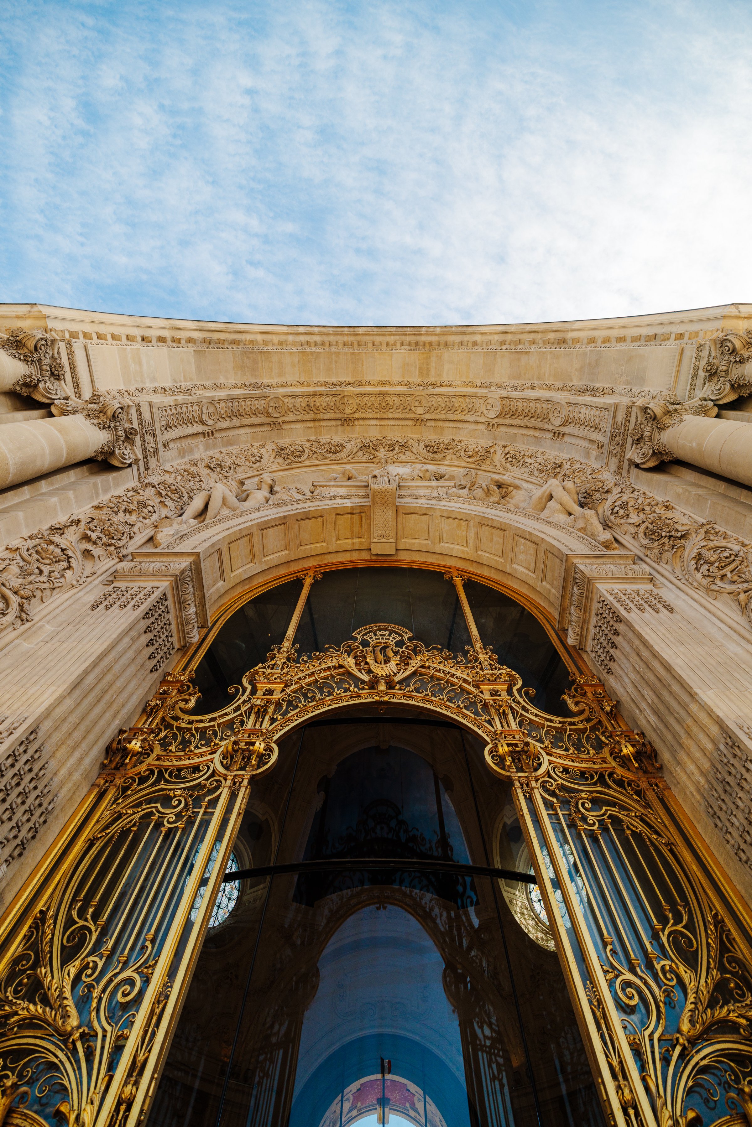 Close-up view of an ornate, gold-accented wrought iron gate at the entrance of an elegant building with detailed stonework and sculptures, viewed from below against a blue sky with scattered clouds.