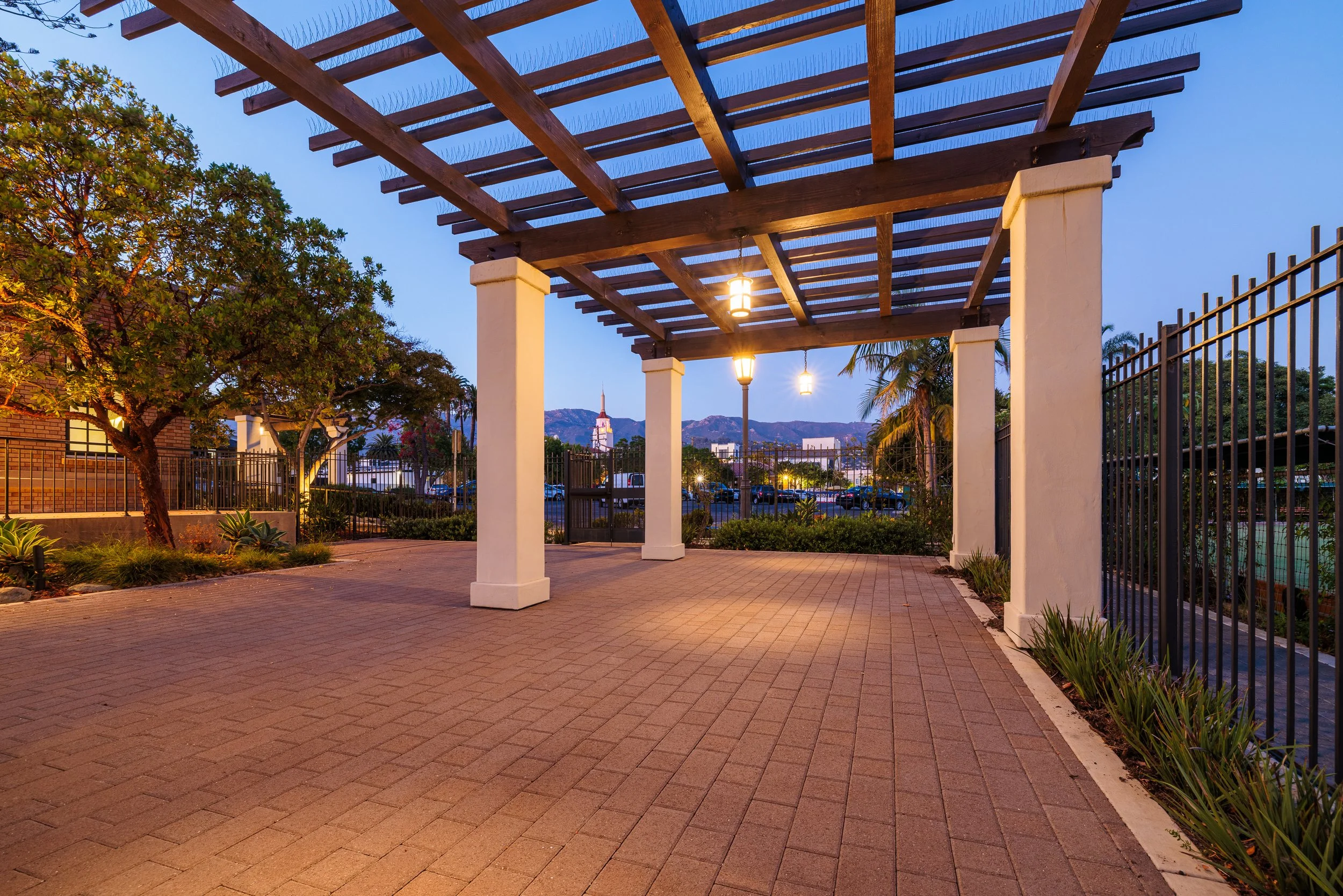 An outdoor courtyard with a wooden pergola, brick paver ground, trees, and landscaping, with mountains, a cityscape, and a church steeple illuminated in the background at dusk.