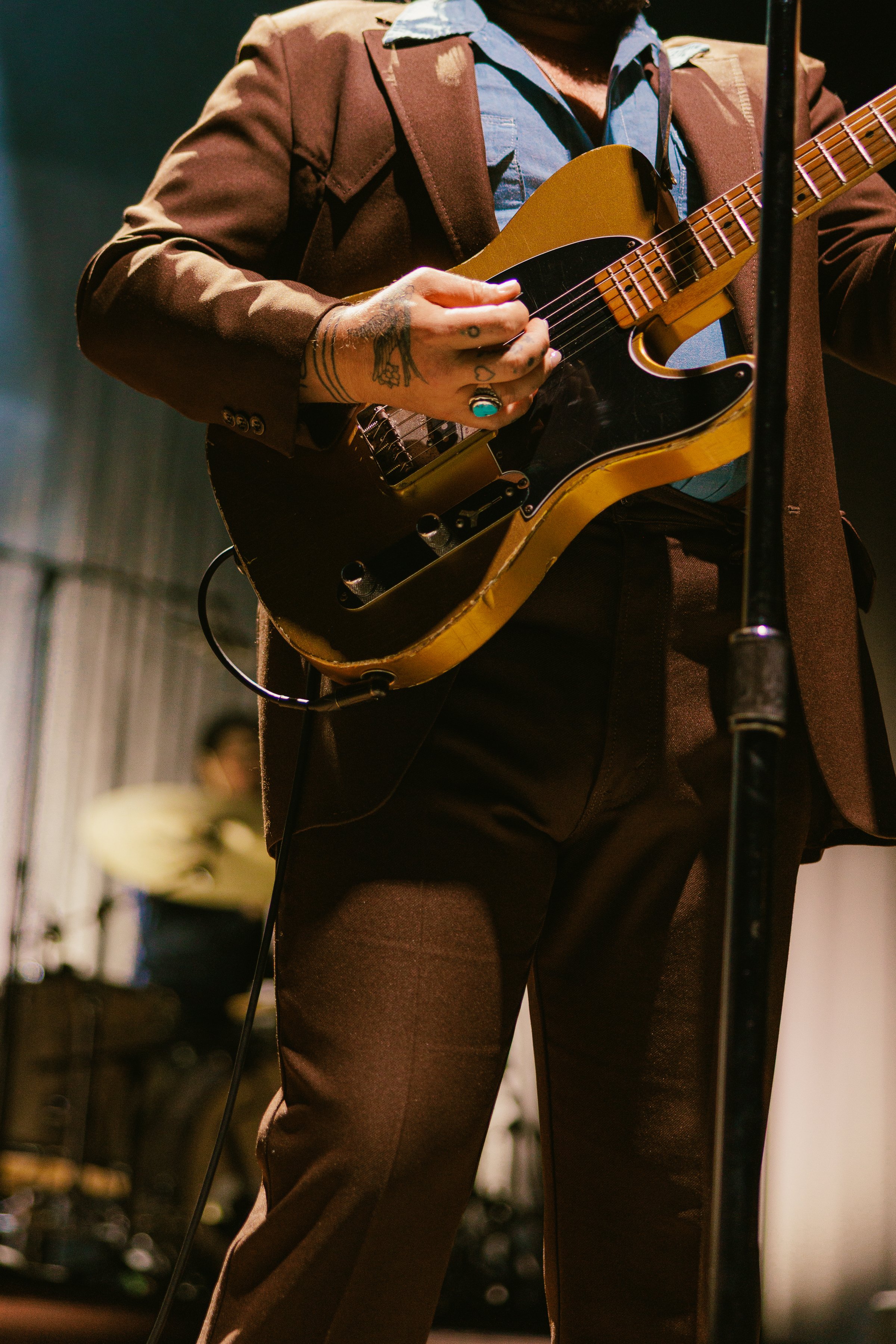 A musician wearing a brown suit playing a vintage yellow and black electric guitar on stage, with a blurred drummer in the background.