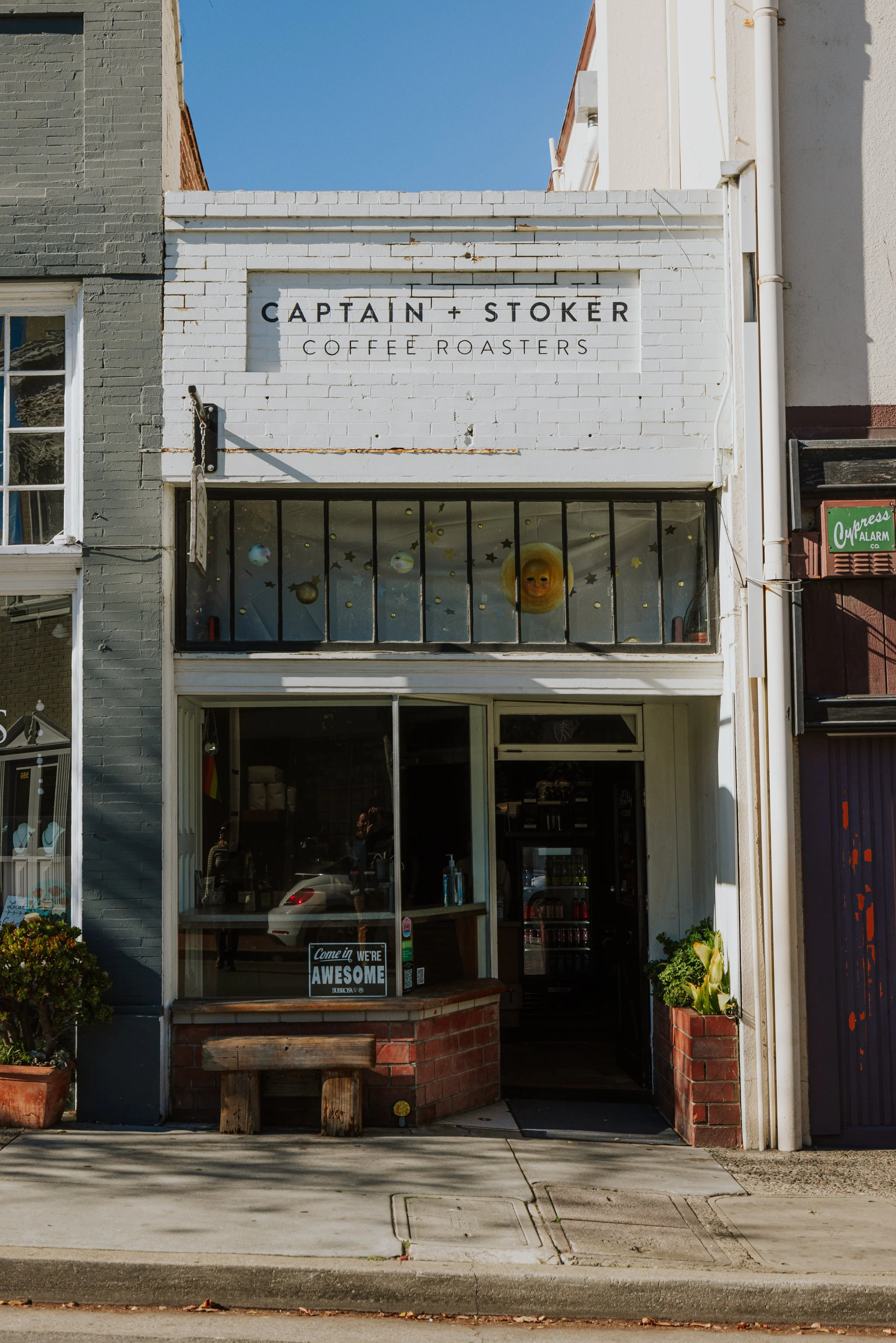 Facade of Captain + Stoker Coffee Roasters with a brick bench outside, window decorations featuring stars and a sun face, and a sign that reads 'Come in we're awesome'.