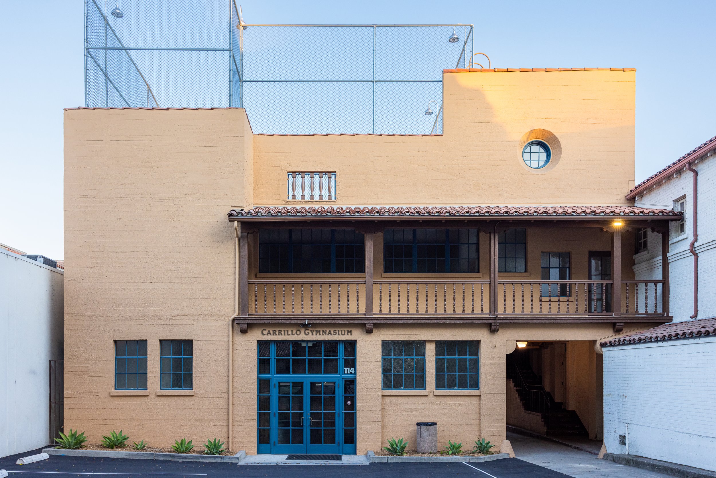 Front view of a two-story building with a yellow facade, blue door, and a wooden balcony on the second floor, labeled 'Carrillo Gymnasium,' with a parking lot in front and a small landscaped area with plants.