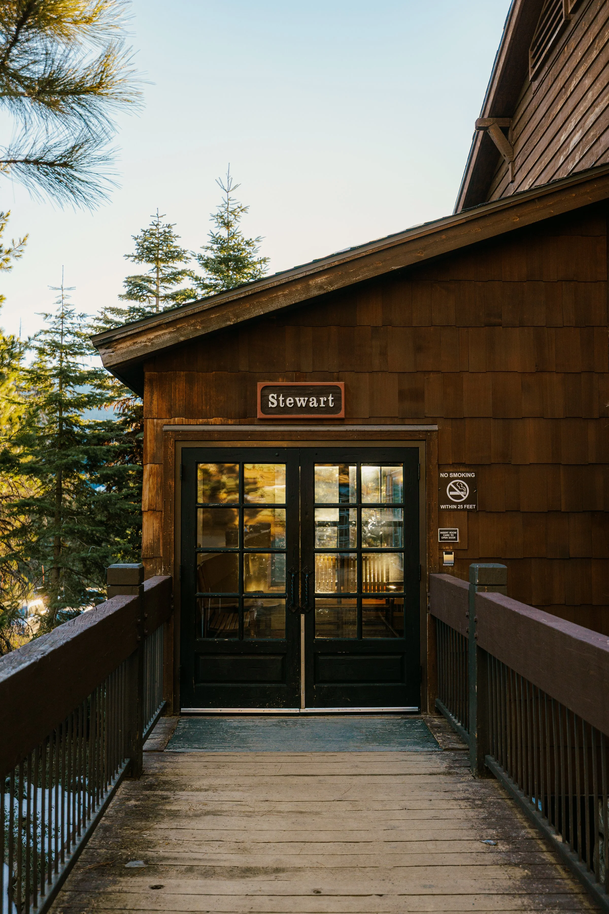 A wooden building entrance with a sign reading 'Stewart', surrounded by trees, with glass double doors and a no smoking sign.