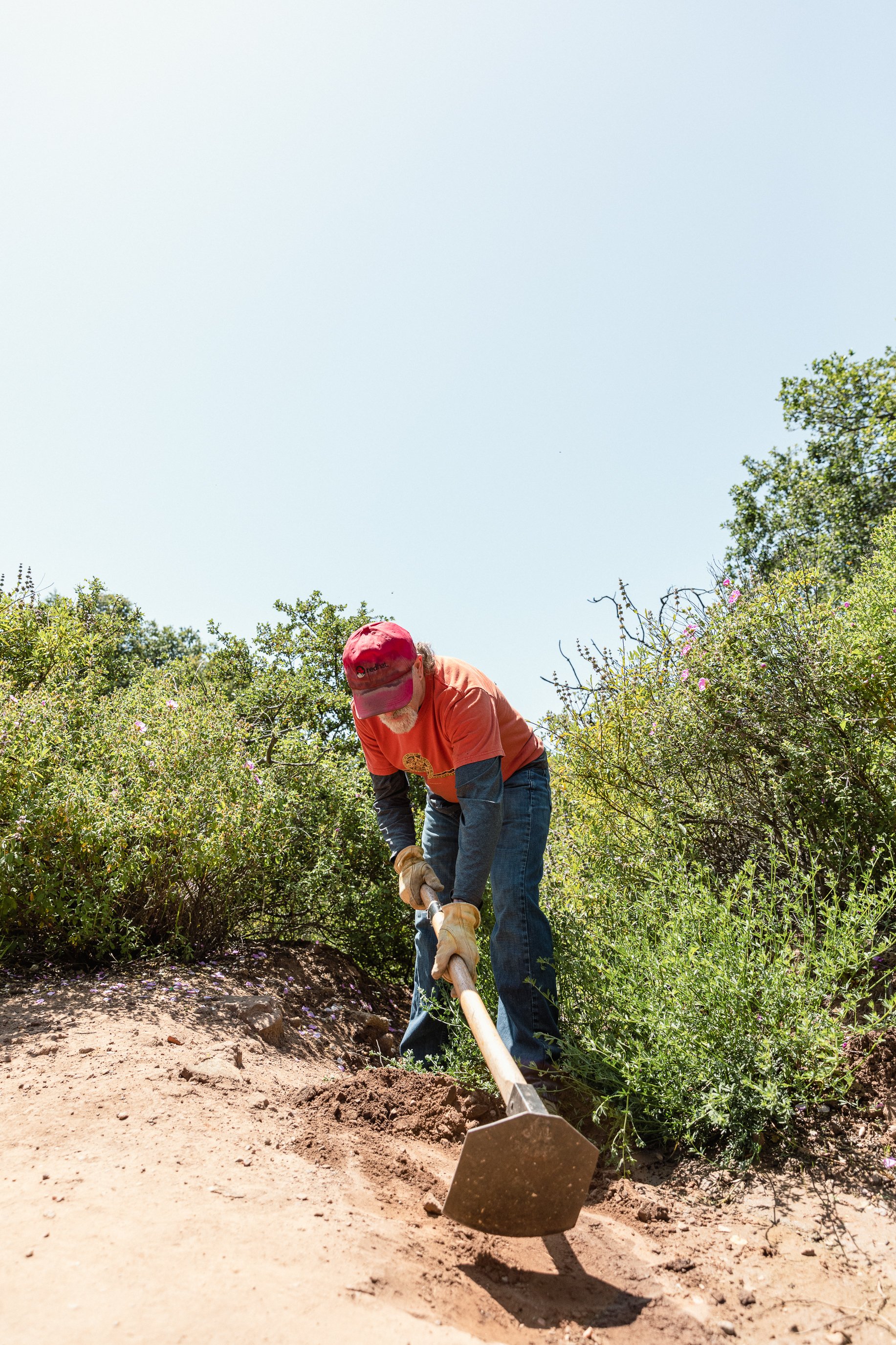 An elderly man wearing a red cap, orange shirt, and gloves digging with a shovel in a sunny outdoor setting surrounded by green bushes and plants.