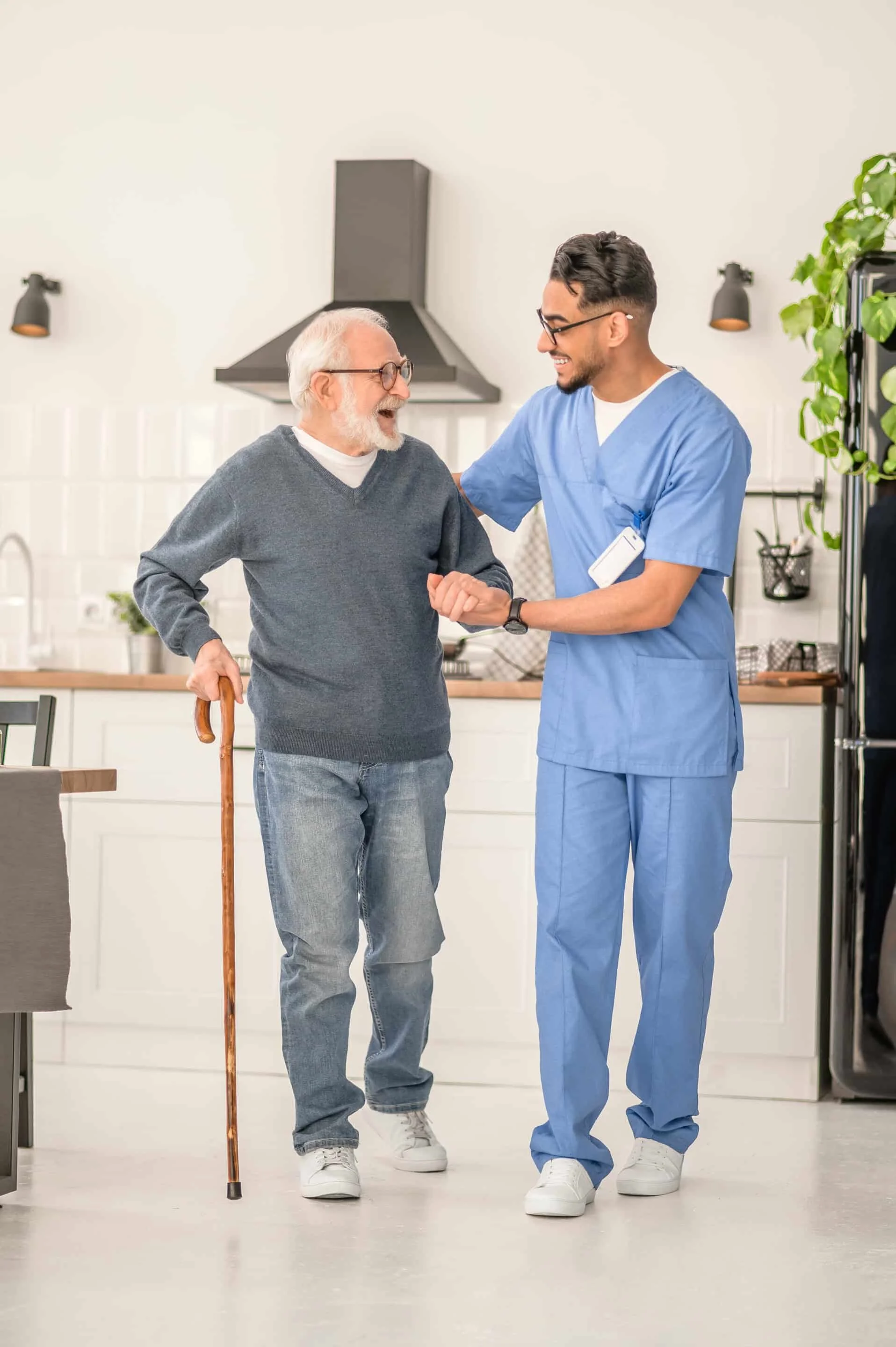A young male nurse in blue scrubs smiling and holding hands with an elderly man with white hair and glasses, who is using a cane, in a bright kitchen.