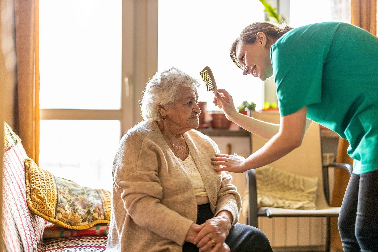 A young woman styling an elderly woman's hair with a brush in a cozy home setting.