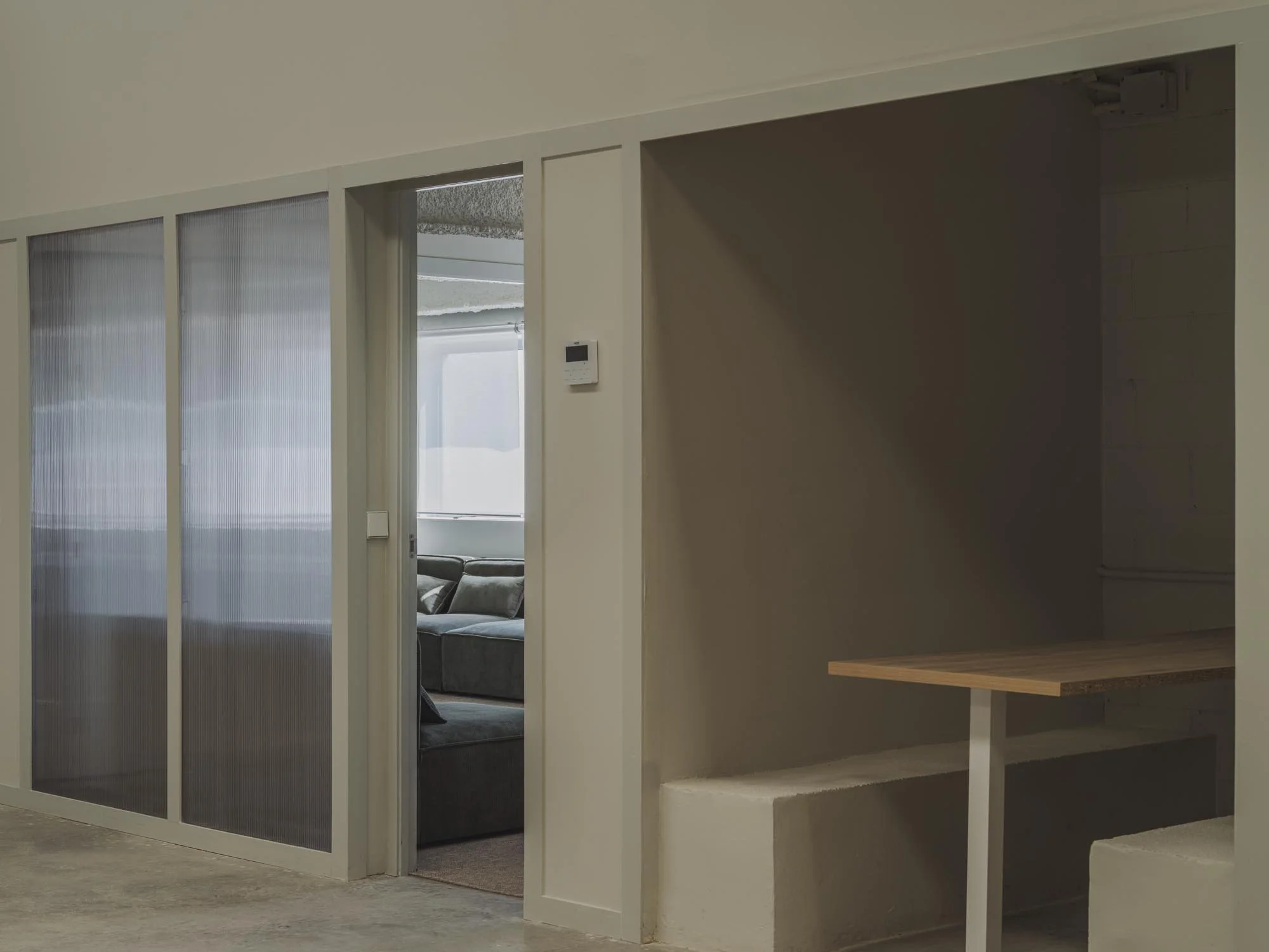 interior de un espacio moderno con pared blanca, un área de descanso con sillones grises y ventanas grandes en el fondo