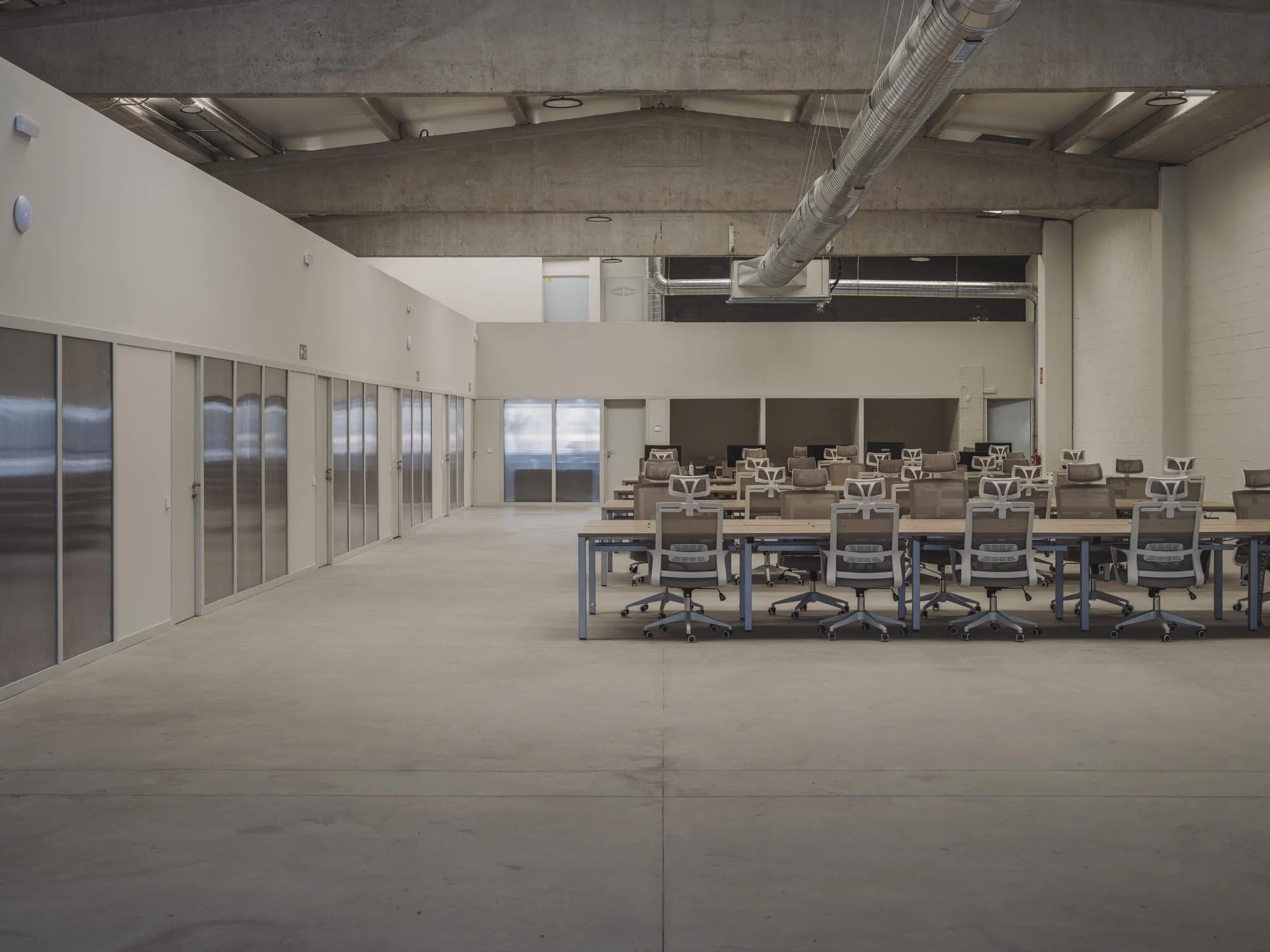 Oficina moderna con mesas y sillas, ventanas grandes, paredes blancas y techo de concreto con ventilación expuesta.