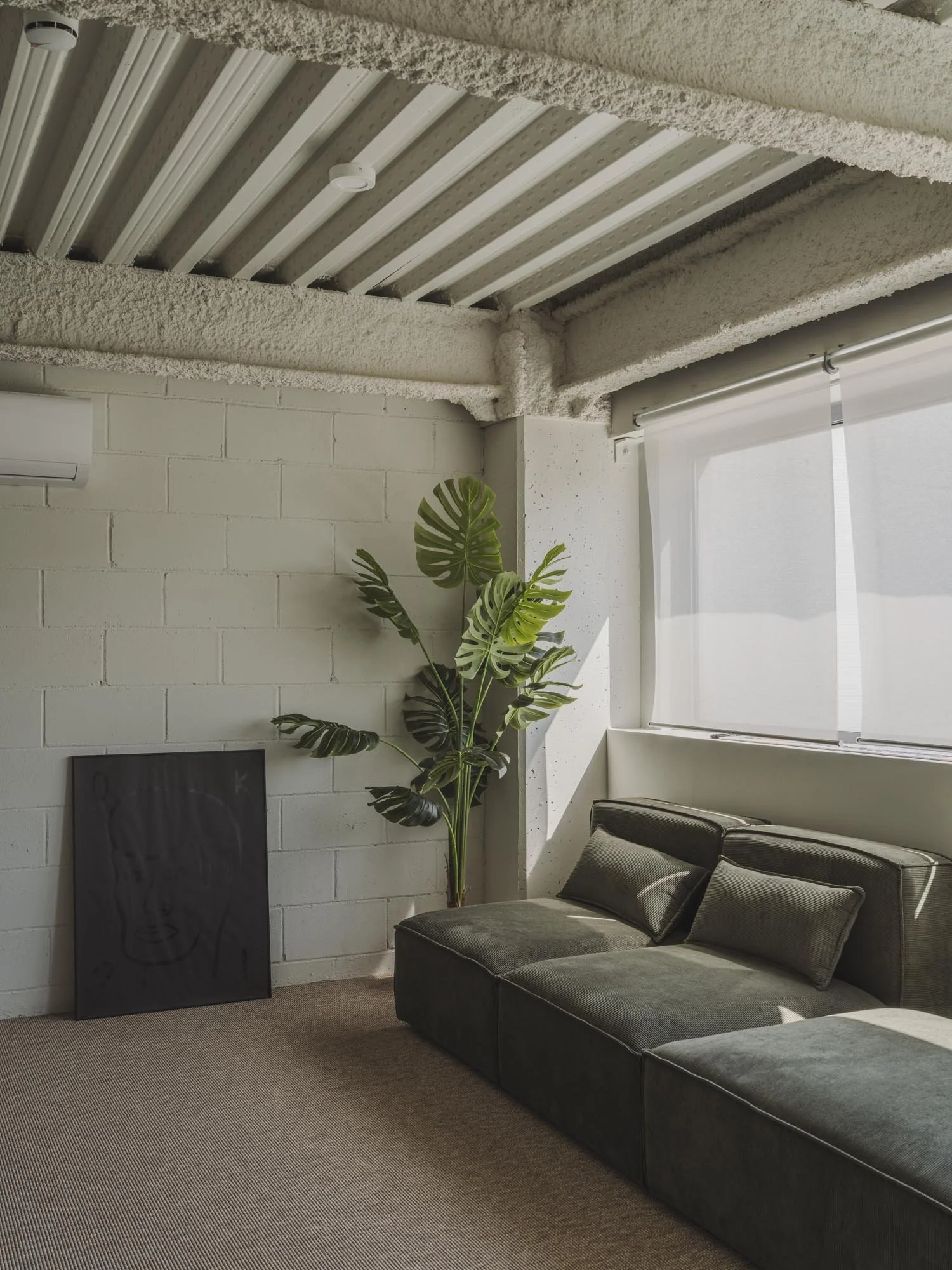 Sala de estar moderna con sofá verde, planta de hojas grandes junto a la pared y cuadro negro apoyado en el suelo, con ventana grande con cortina blanca y muro de bloques de concreto blanco.