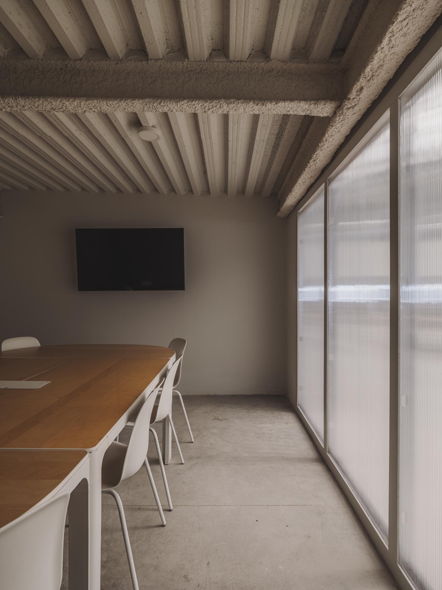 Sala de reuniones con mesa de madera blanca y sillas blancas, pared gris claro, pantalla de televisión negra en la pared, ventanas grandes de cristal con cortinas translúcidas, techo de concreto con vigas expuestas.