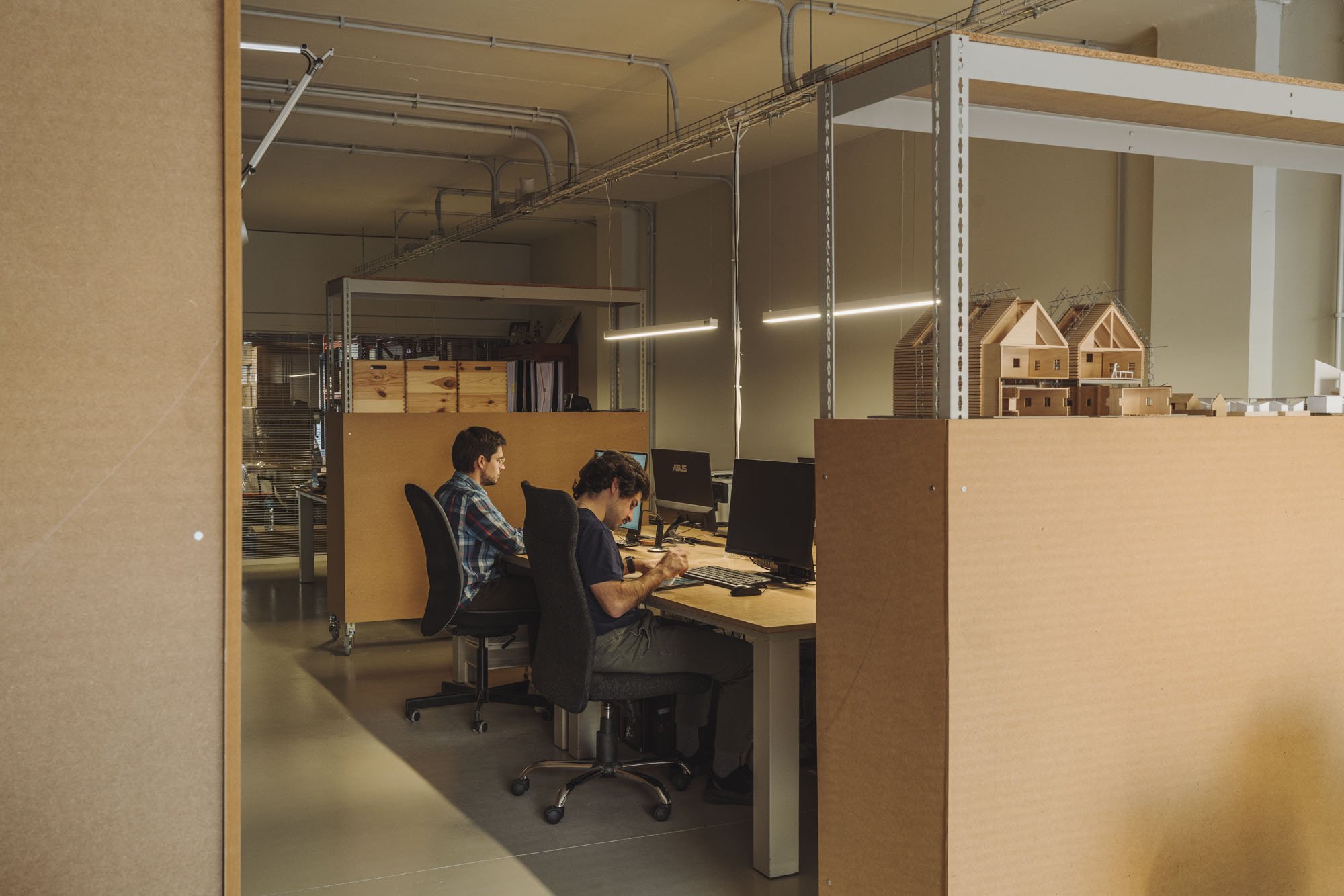 Área de trabajo con dos personas en computadoras, muebles de madera y modelos de casas en miniatura en una oficina moderna.