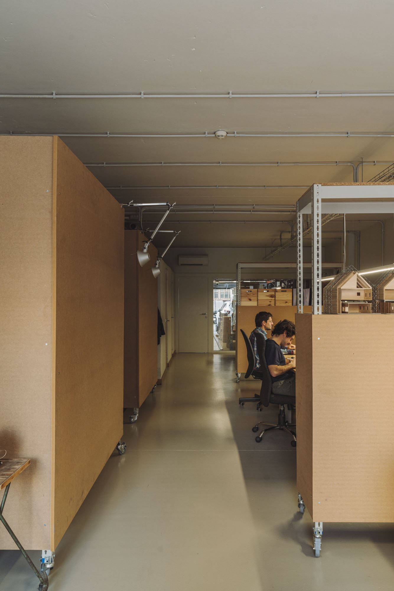Espacio de trabajo con personas sentadas en escritorios, estructuras de madera, y estantes metálicos, en un ambiente moderno y organizado.