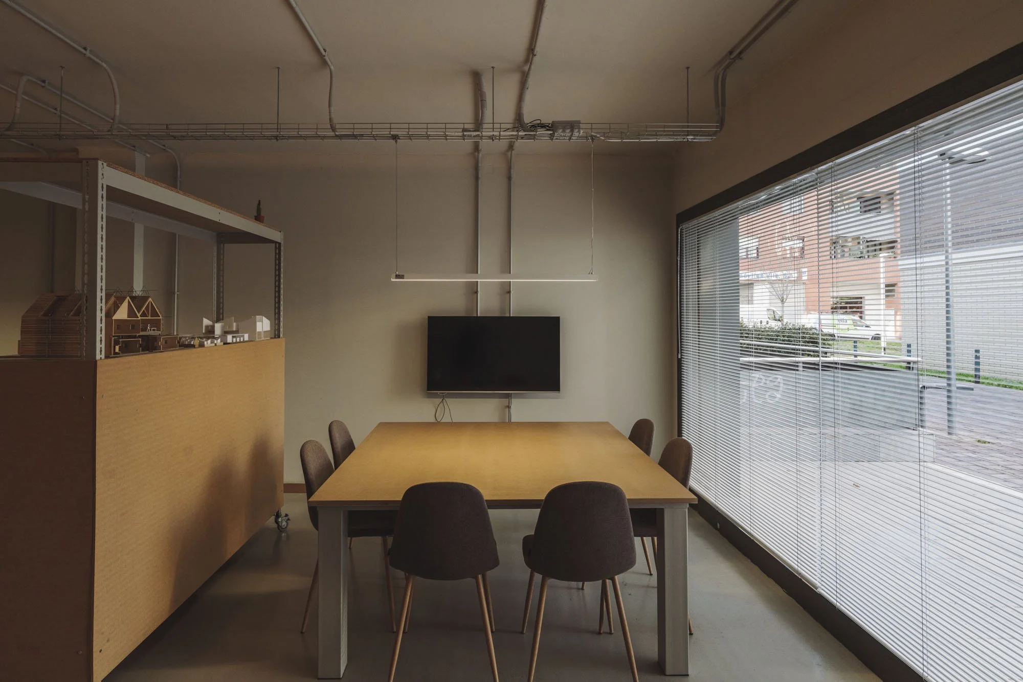 Sala de reuniones con mesa de madera y seis sillas tapizadas, pantalla plana en la pared, ventanas con persianas horizontales, y modelos arquitectónicos sobre una estantería.
