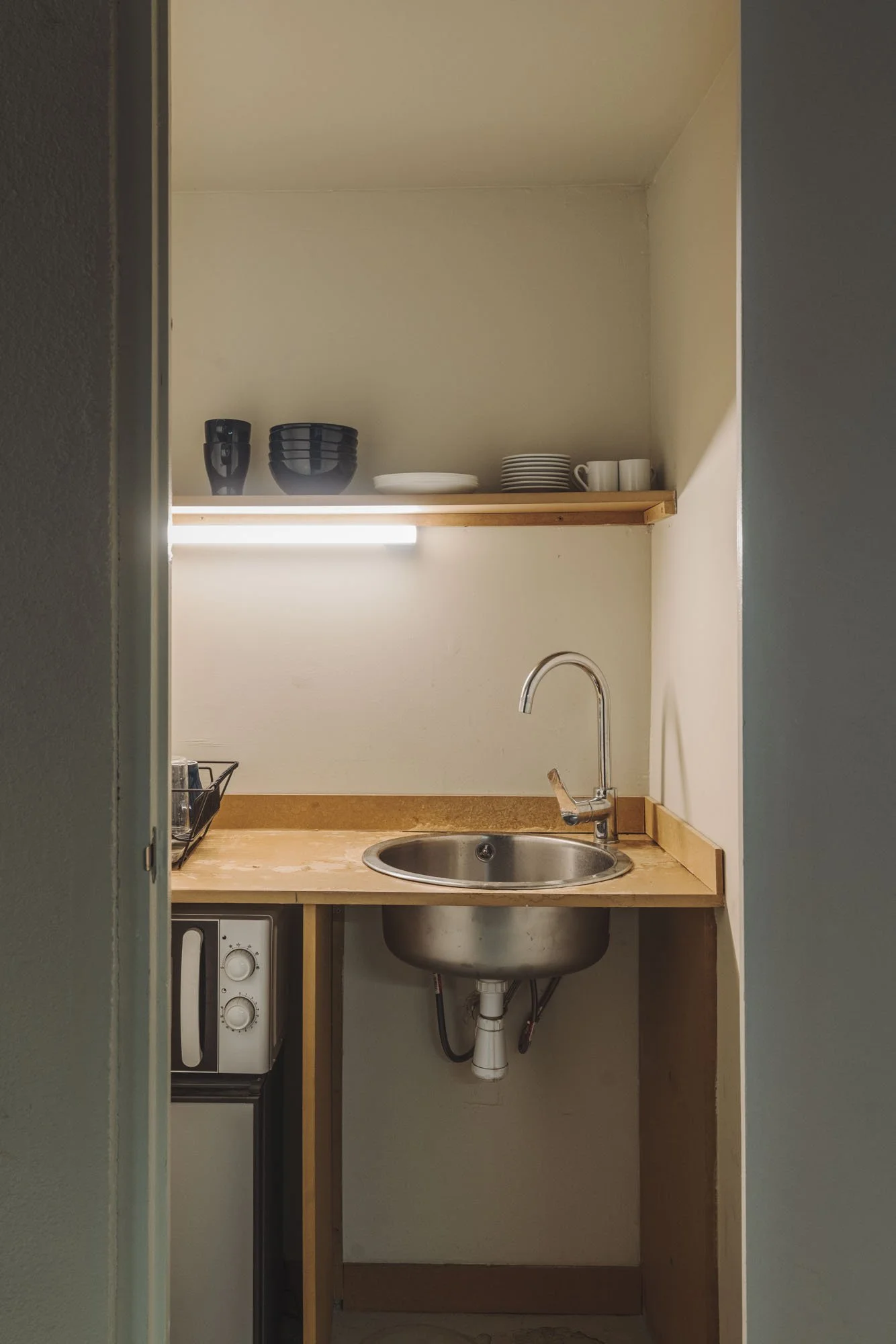 Pequeña cocina con lavabo de acero inoxidable, estantes con platos y tazas, microondas, y espacio de trabajo de madera.