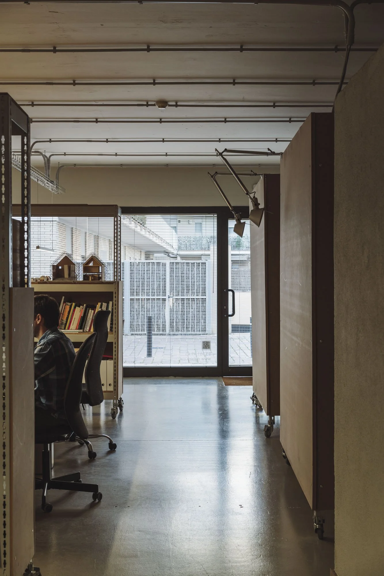 Interior de una oficina con escritorios, sillas, estantes con libros y objetos decorativos, y una puerta de vidrio que da a una zona exterior con piso de azulejo y rejas metálicas.