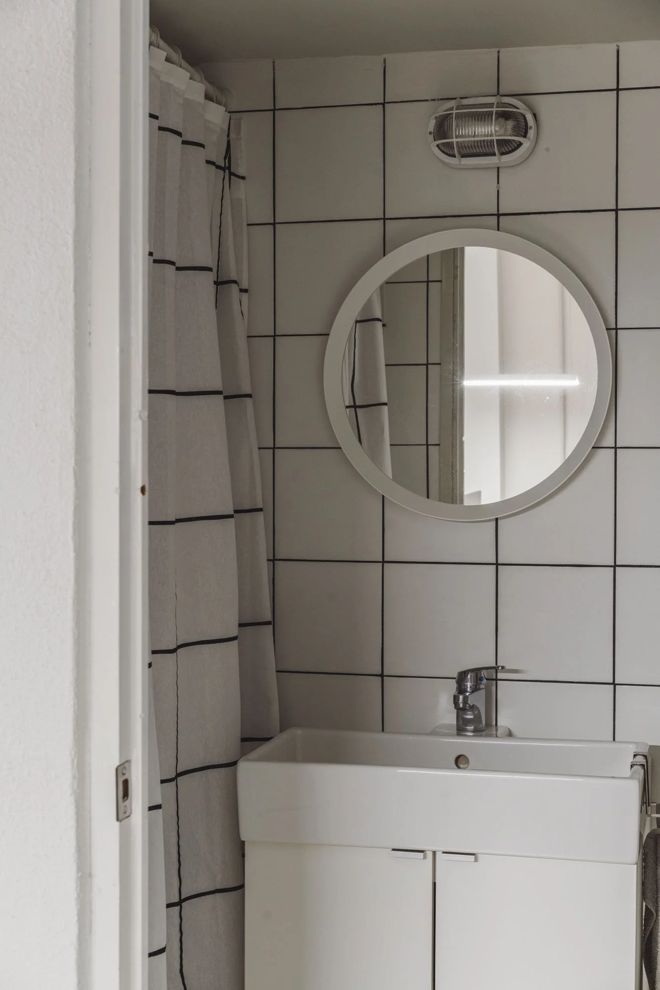 Vestidor de baño con azulejos blancos, espejo circular, lavabo con mueble blanco, cortina de ducha a rayas blancas y negras y extractor de aire en la pared.