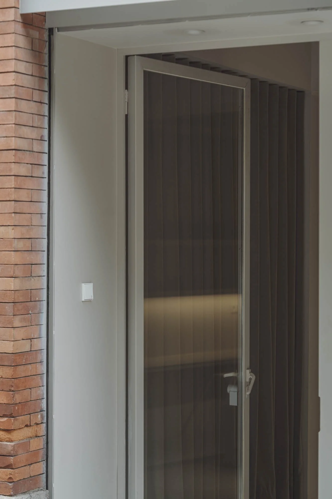 Puerta de vidrio con cortinas oscuras en el interior, marco de aluminio, paredes blancas y muro de ladrillos a la izquierda.