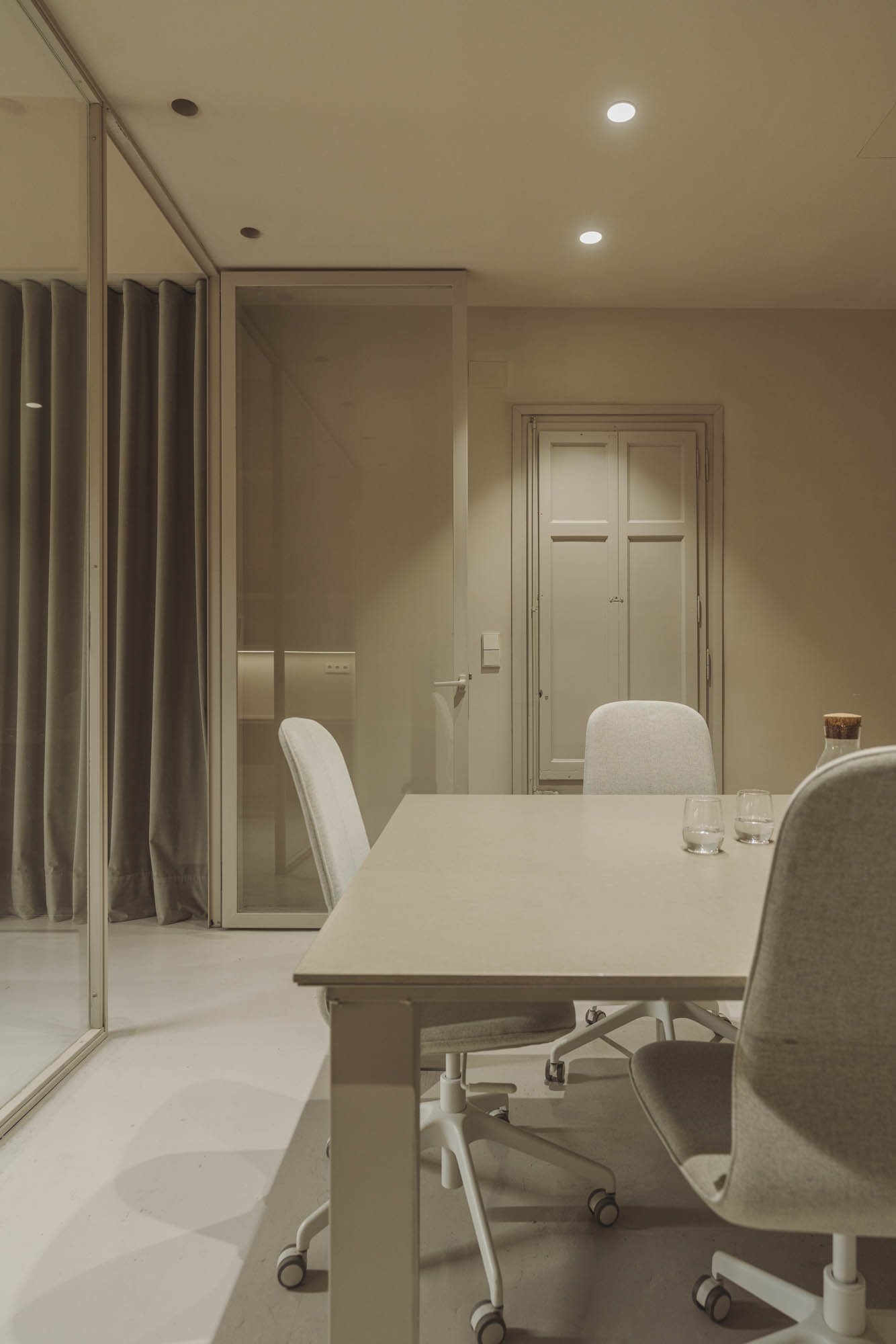 Sala de reuniones vacía con una mesa blanca y sillas grises, una pared de vidrio y cortinas, puerta cerrada y techo con luces empotradas.