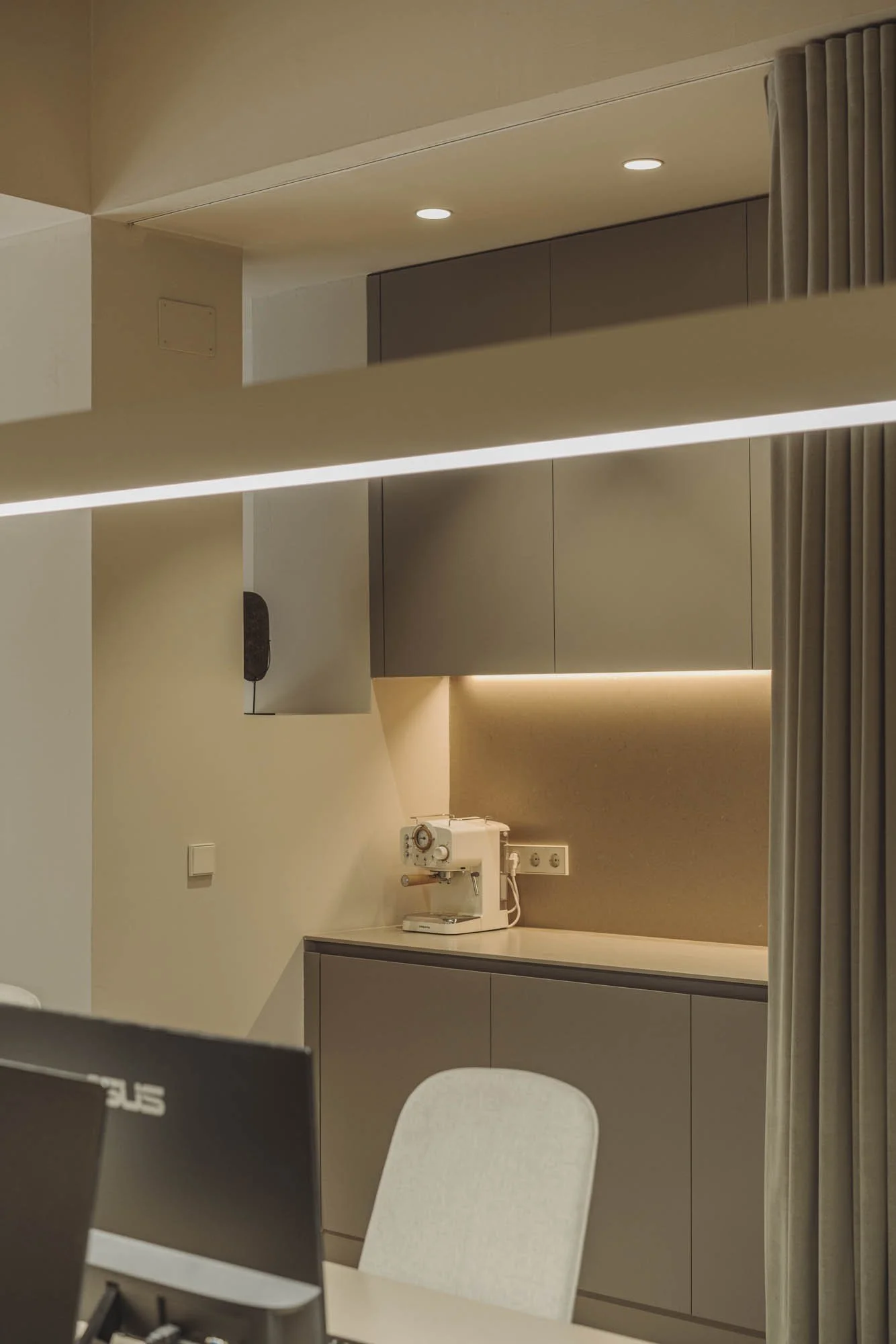 Vista parcial de una cocina moderna con gabinetes beige, una máquina de café en la encimera y cortinas beige a la derecha, con iluminación suave y paredes en tonos neutros.
