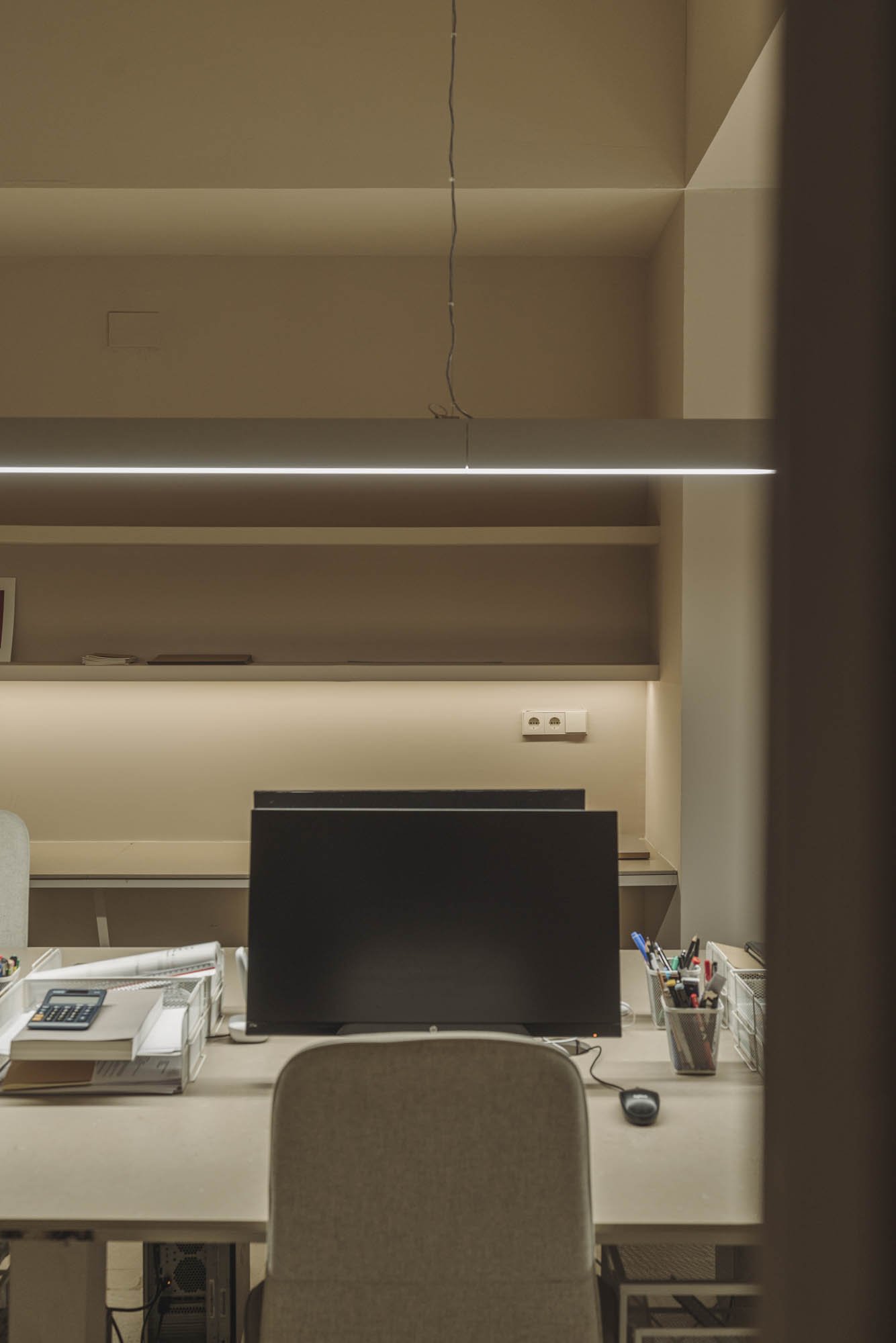 Oficina con escritorio, monitor, ratón, documentos, bolígrafos, toma corriente y estantes de paredes en fondo con iluminación y paredes beige.