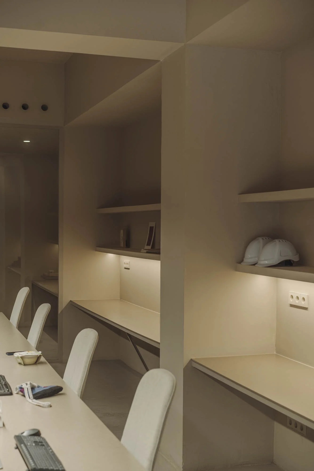 Sala de reuniones con mesa y sillas blancas, estanterías con cascos de seguridad blancos y enchufes en la pared.