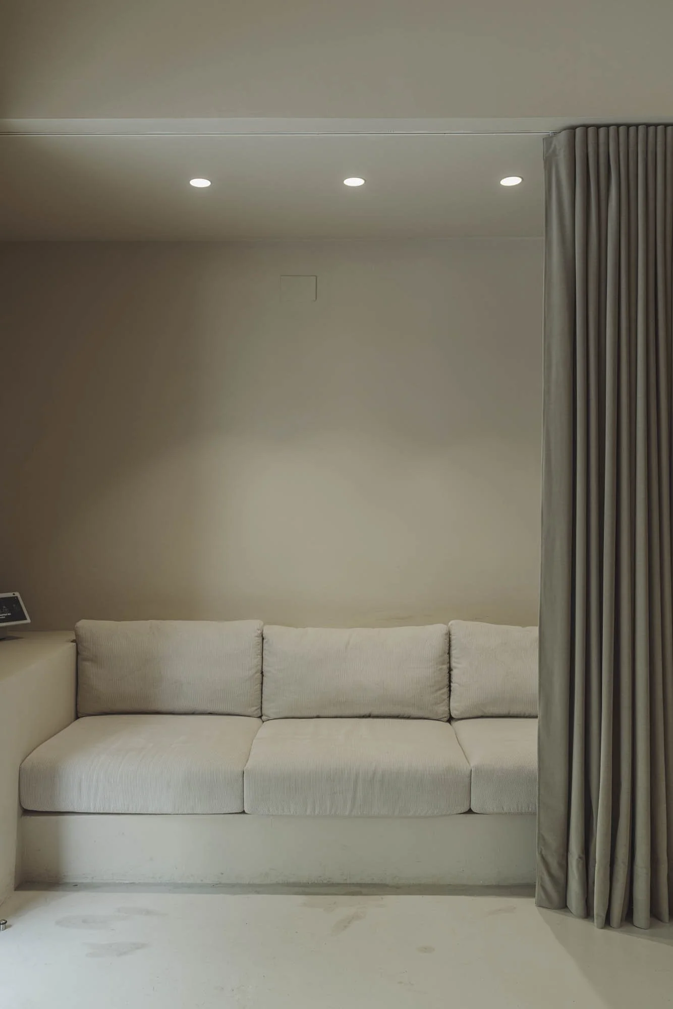 Sofá beige de tres plazas frente a una pared gris y cortinas grisáceas en una sala moderna con iluminación empotrada en el techo.