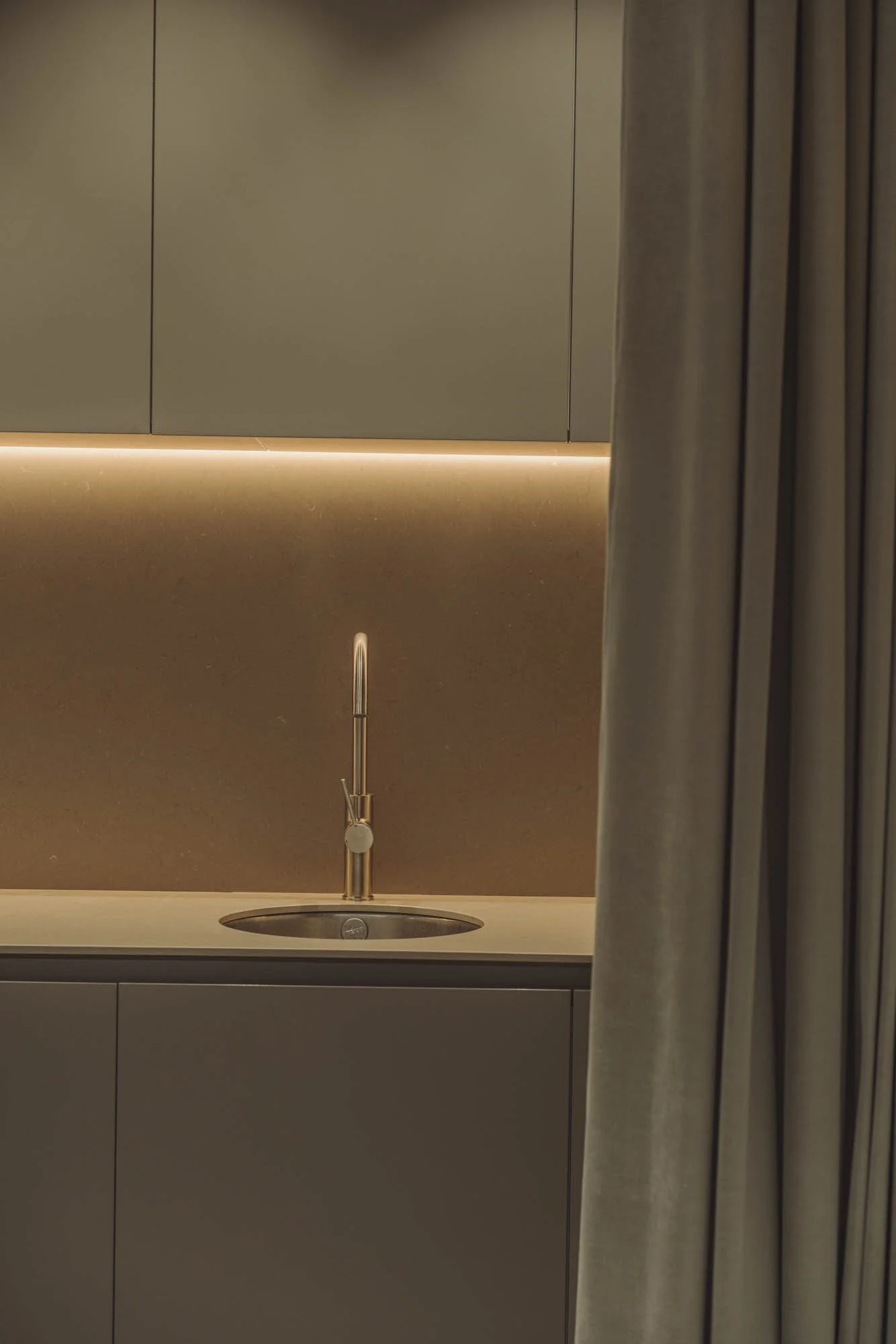 Vista partial de una cocina moderna con fregadero y grifo de metal, cortinas beige en un lado, en tonos cálidos y minimalistas.