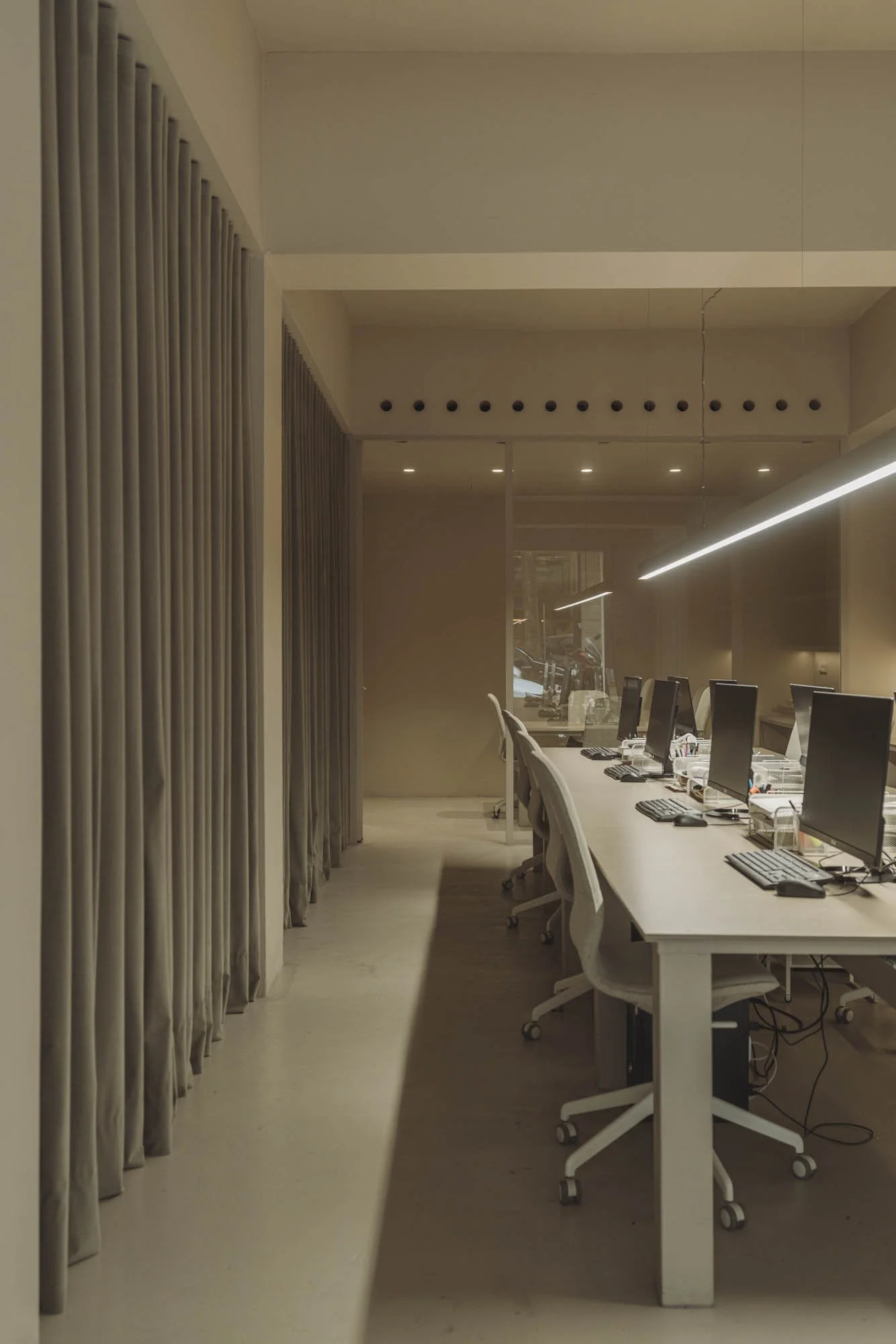 Oficina con computadoras en una mesa larga, sillas blancas y cortinas grises, iluminación moderna y ventana con vista a la calle.
