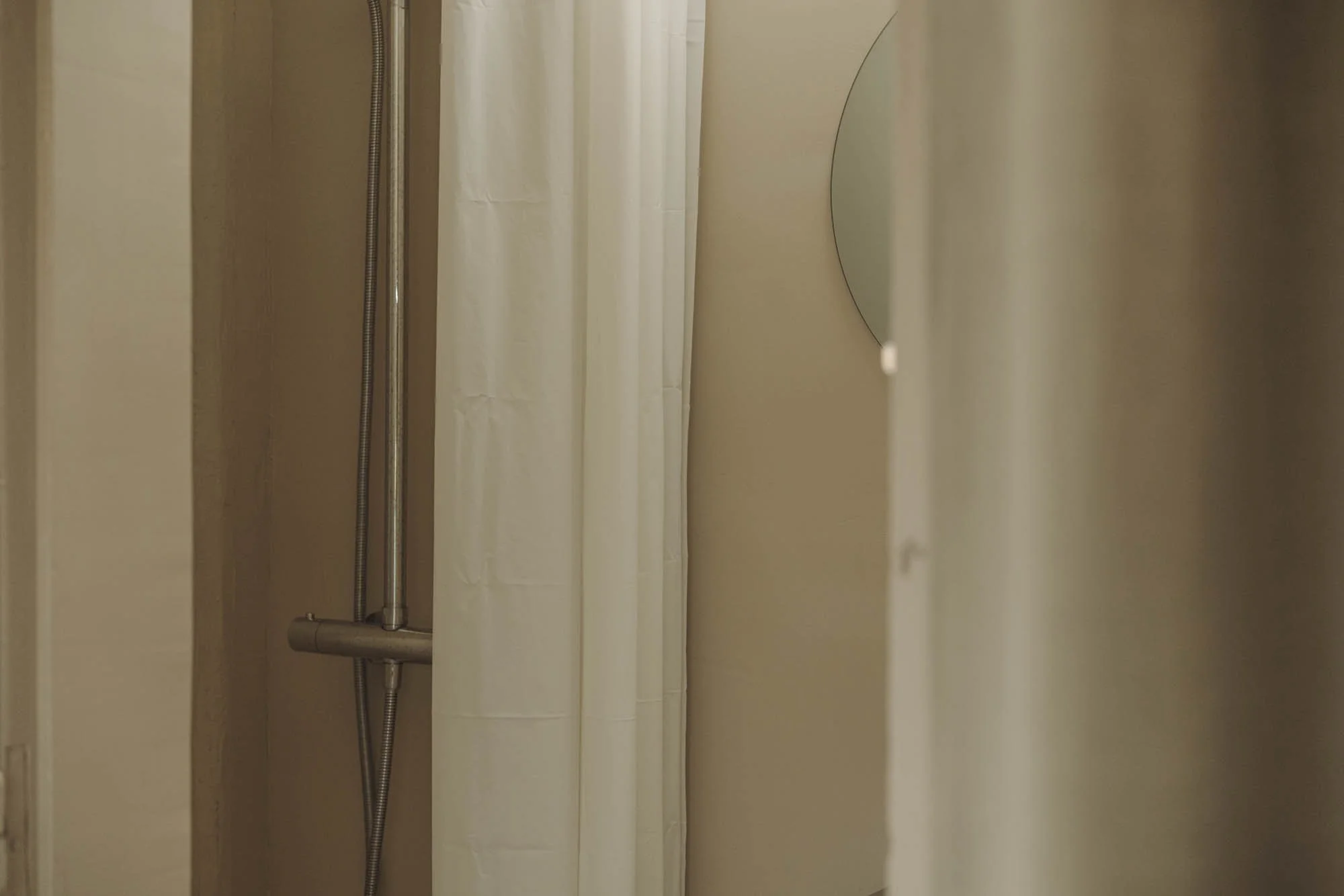 Un espacio de ducha con cortina blanca y un espejo redondo en la pared beige.
