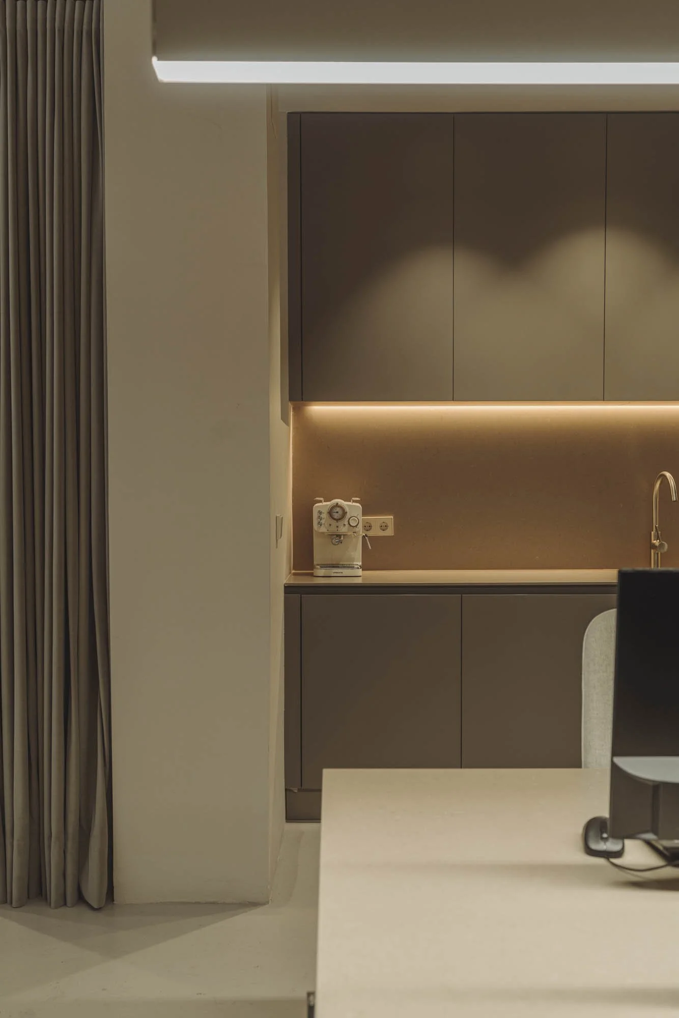 Una cocina moderna con gabinetes grises y encimera con iluminación empotrada, una cafetera antigua en la encimera y un fregadero dorado, con una cortina beige a la izquierda y un escritorio con computadora en primer plano.